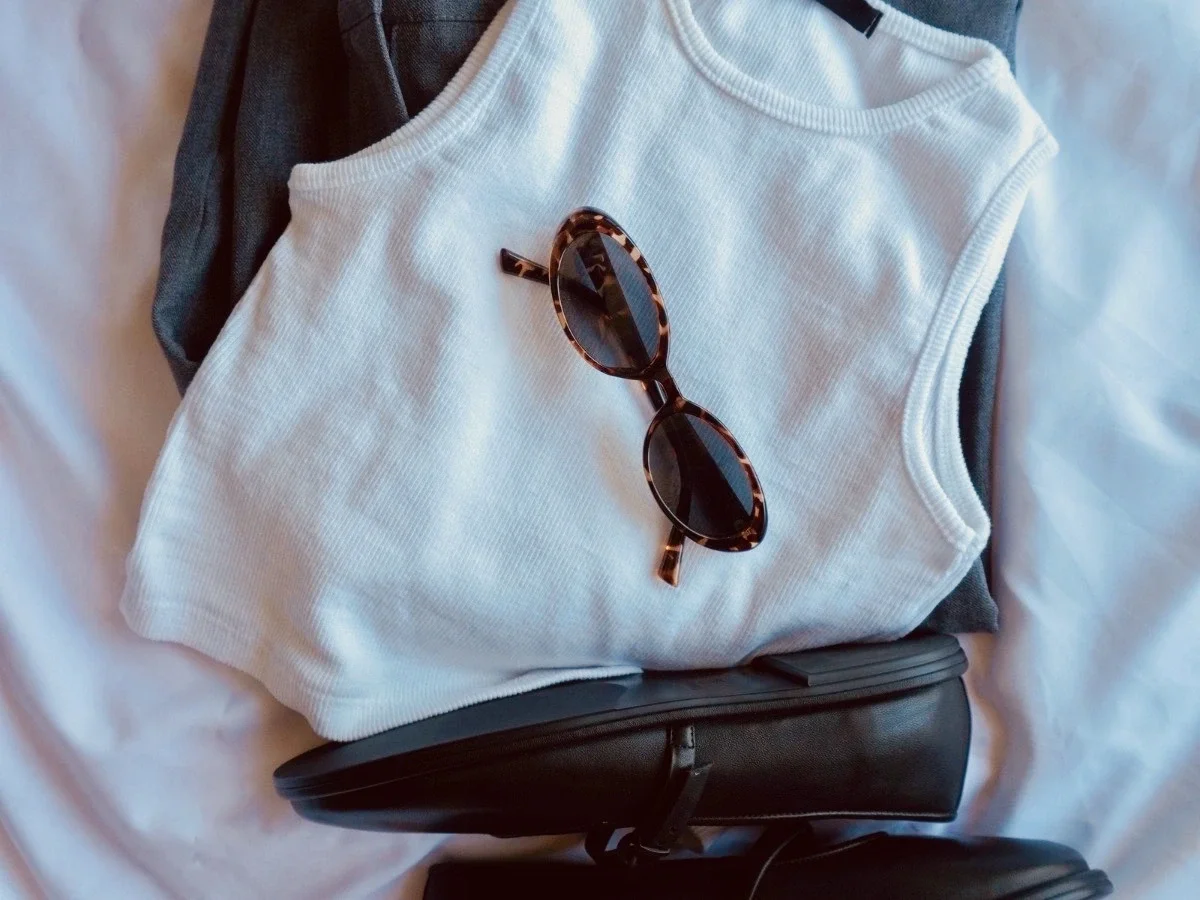 Minimalist flat lay of a white ribbed tank top, black loafers, and tortoiseshell sunglasses on a bed, representing a curated wardrobe audit