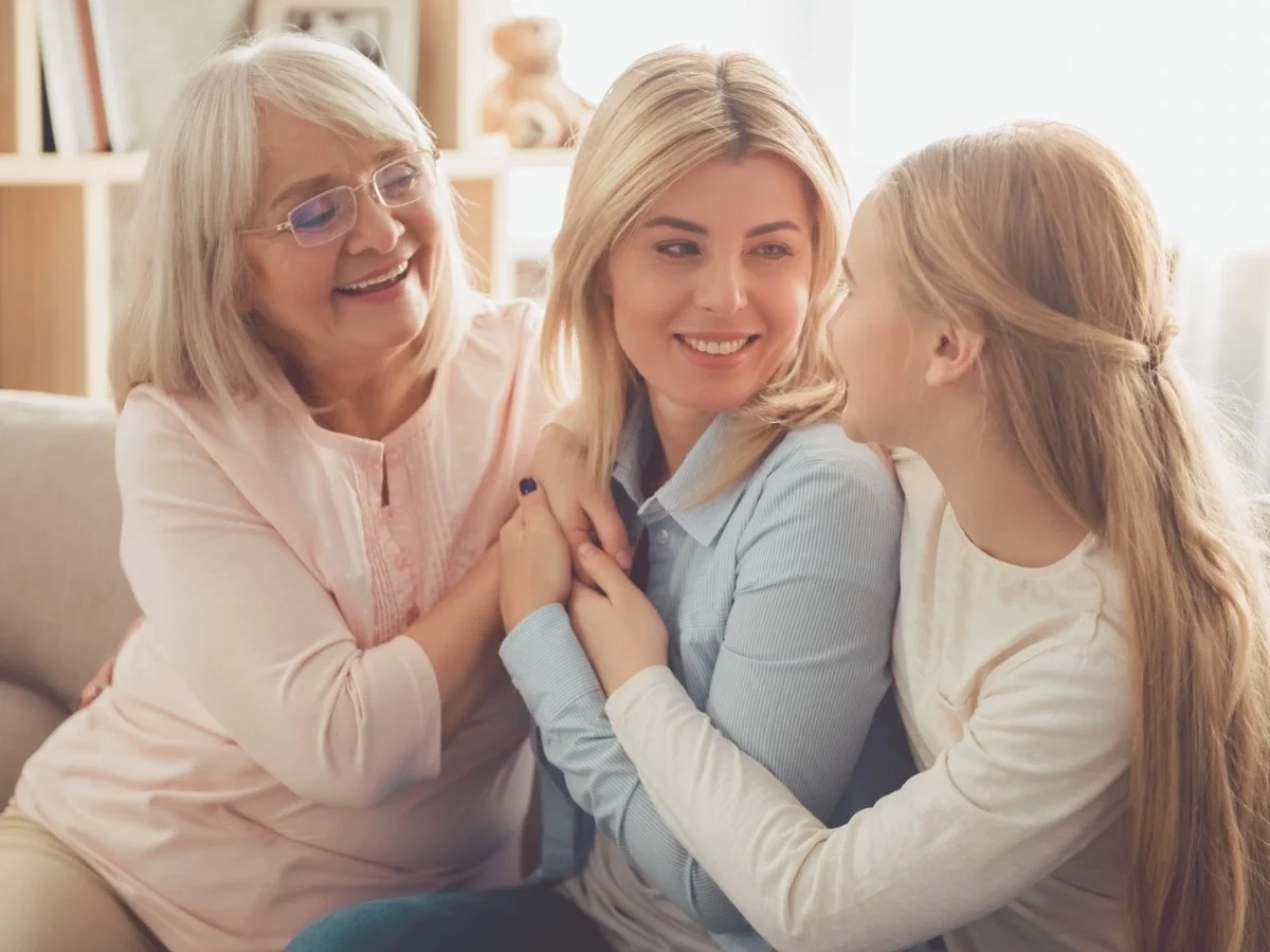 Three generations of women — grandmother, mother, and daughter — sitting closely together smiling, representing the warmth and expressiveness that coexists with generational silence across cultures.