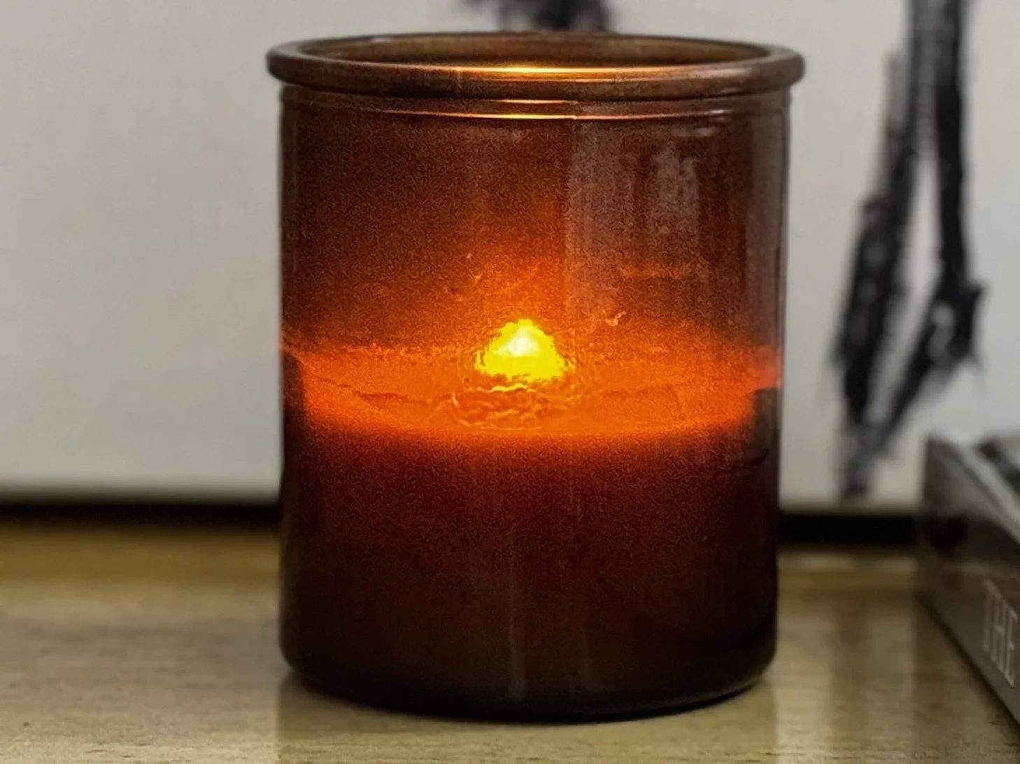 A lit candle glowing amber inside a dark glass vessel, casting warm light against a neutral background