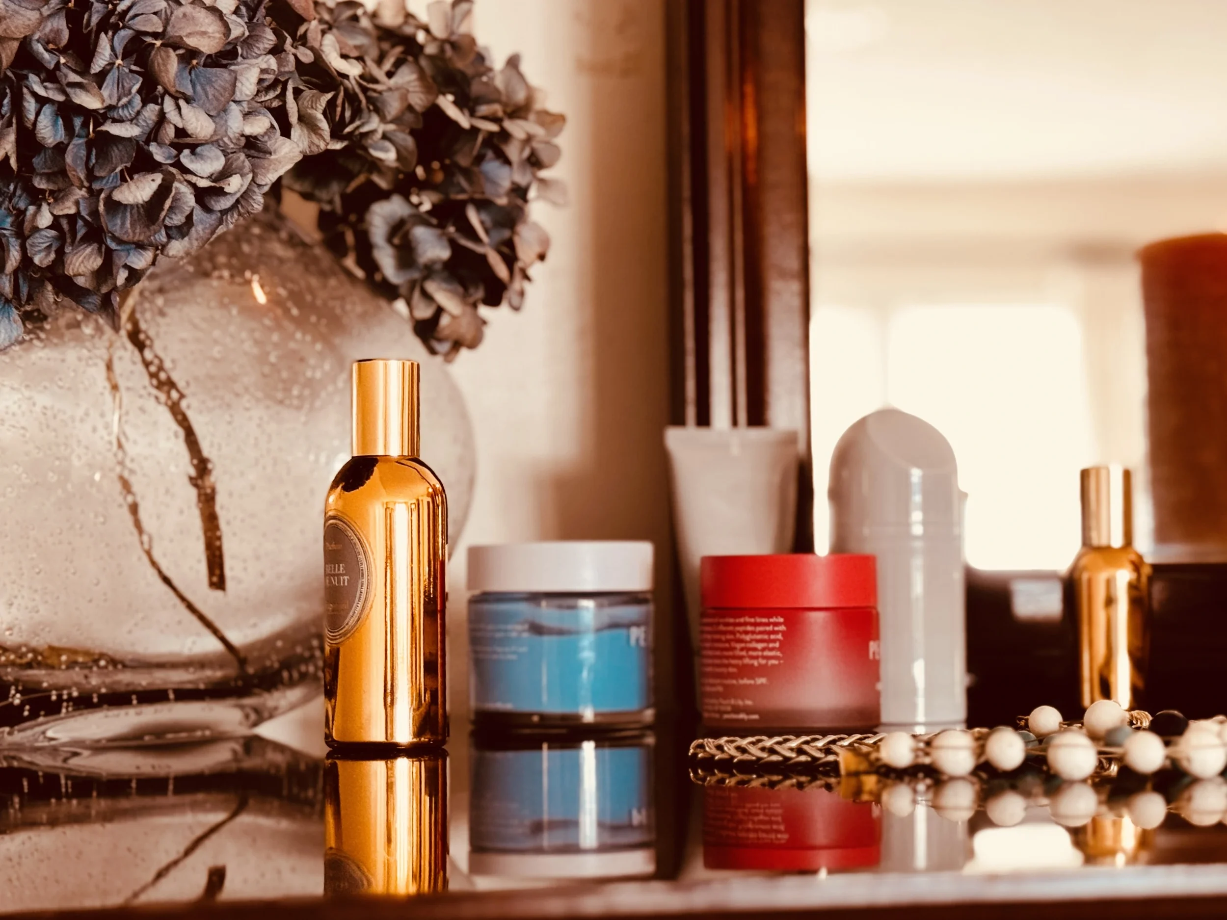 Single gold perfume bottle on a reflective surface with dried blue hydrangeas, representing the intentional process of choosing a signature scent