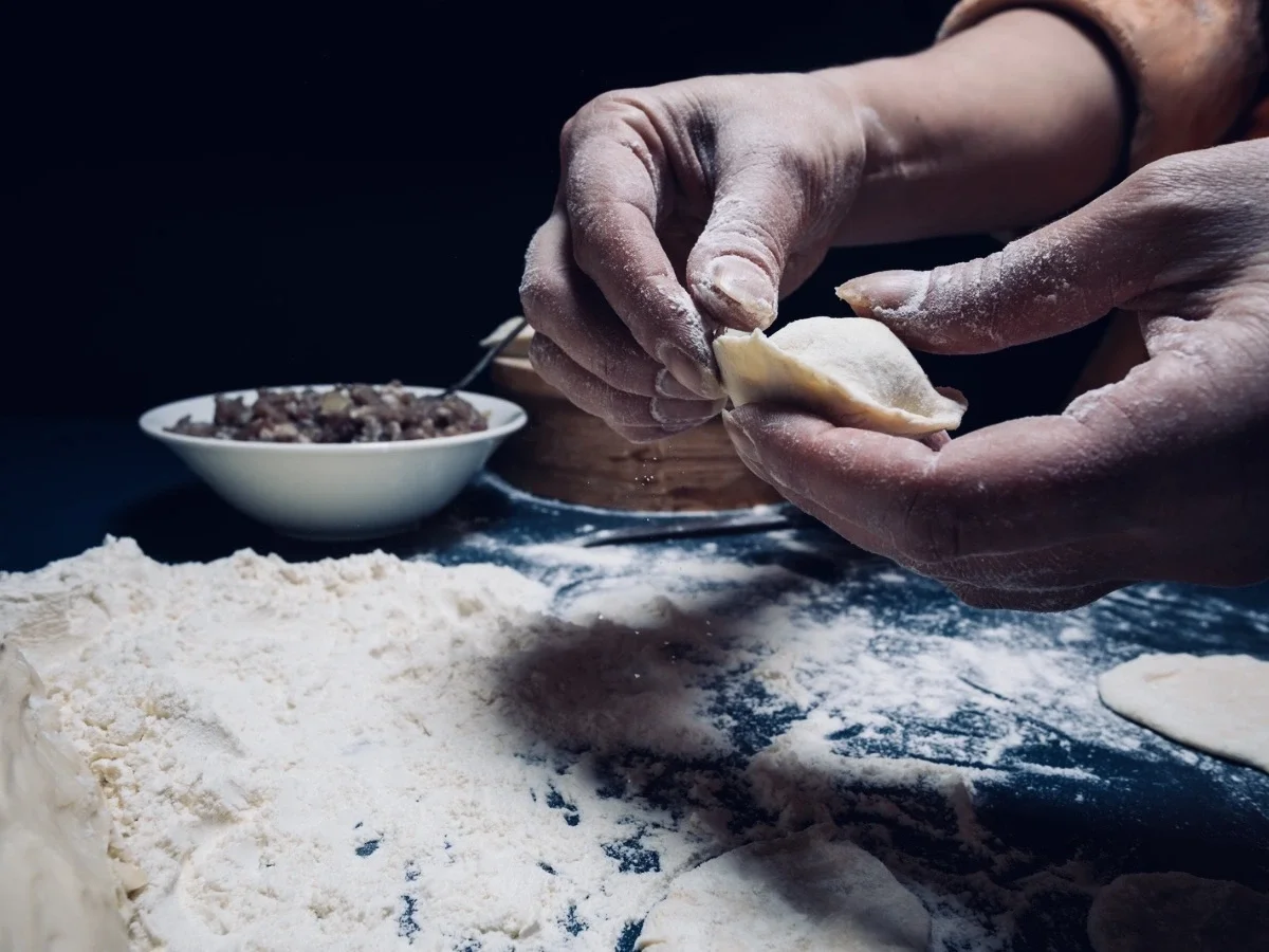 Flour-dusted hands folding a dumpling over a dark surface, evoking silent care passed between generations
