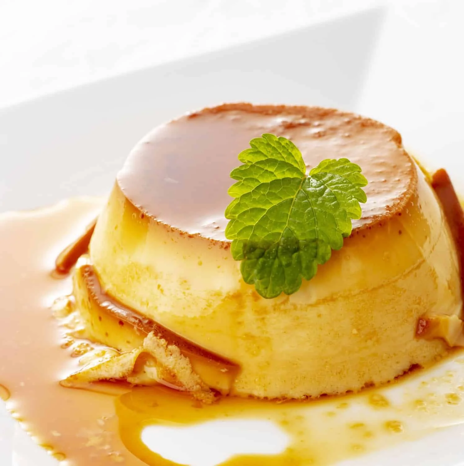 Smooth Cuban flan topped with caramel sauce and a mint leaf on a white plate