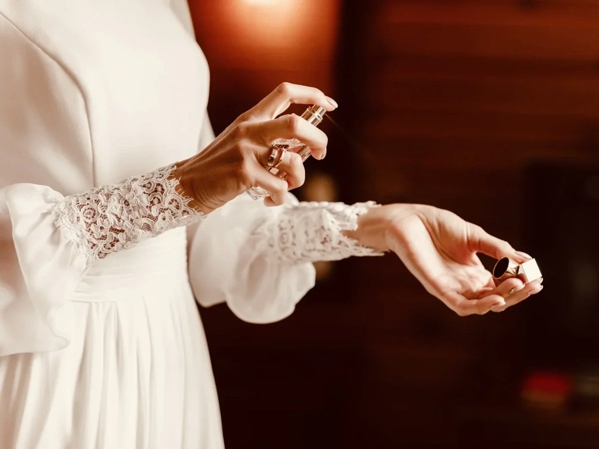 Woman in an elegant white lace-sleeve dress spraying perfume on her wrist, representing the intentional and personal ritual of wearing a signature scent