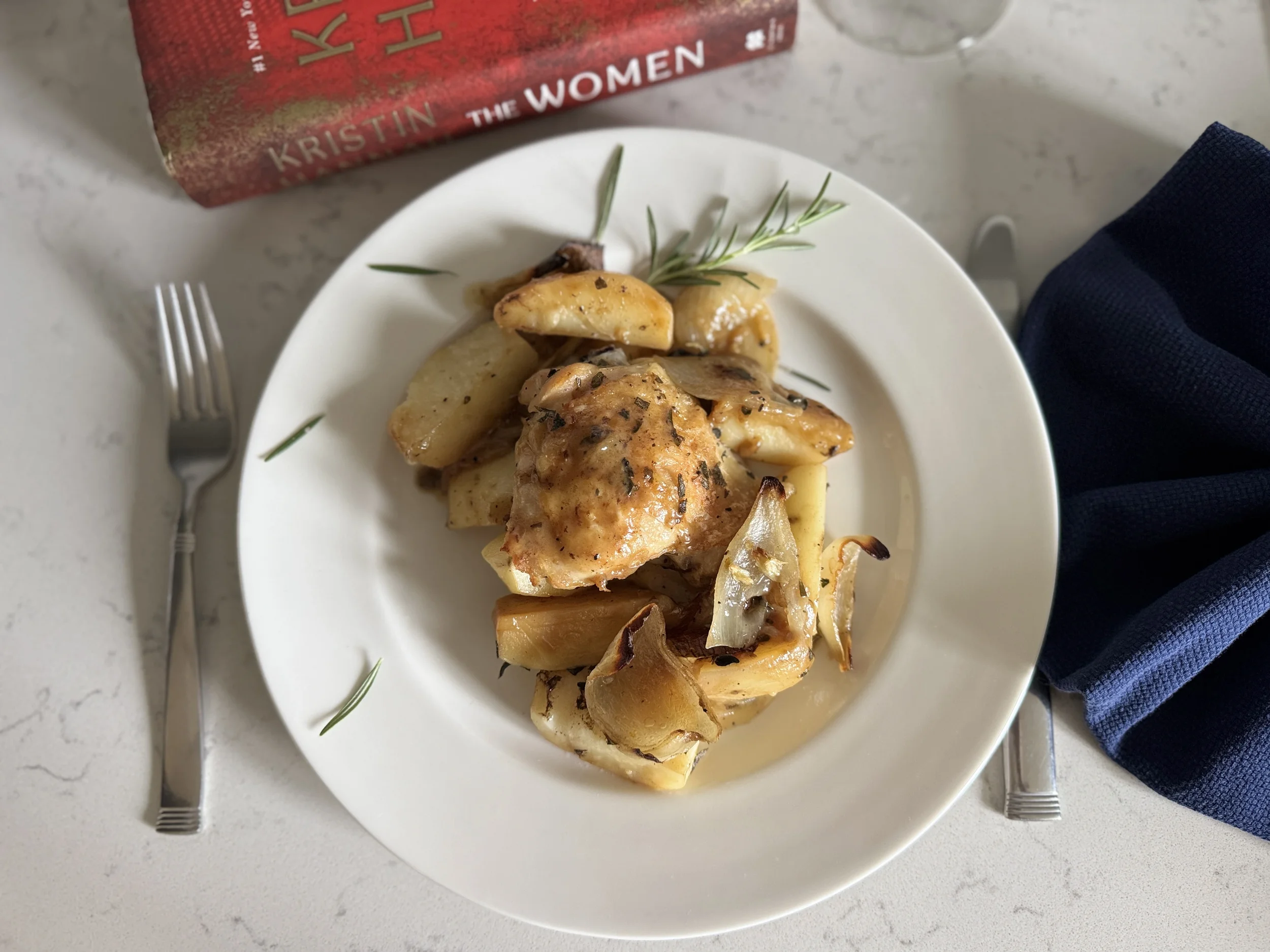 Roasted chicken, potatoes, and onions plate beside Kristin Hannah's novel The Women and a glass of white wine—a dinner pairing for Henry's chapters