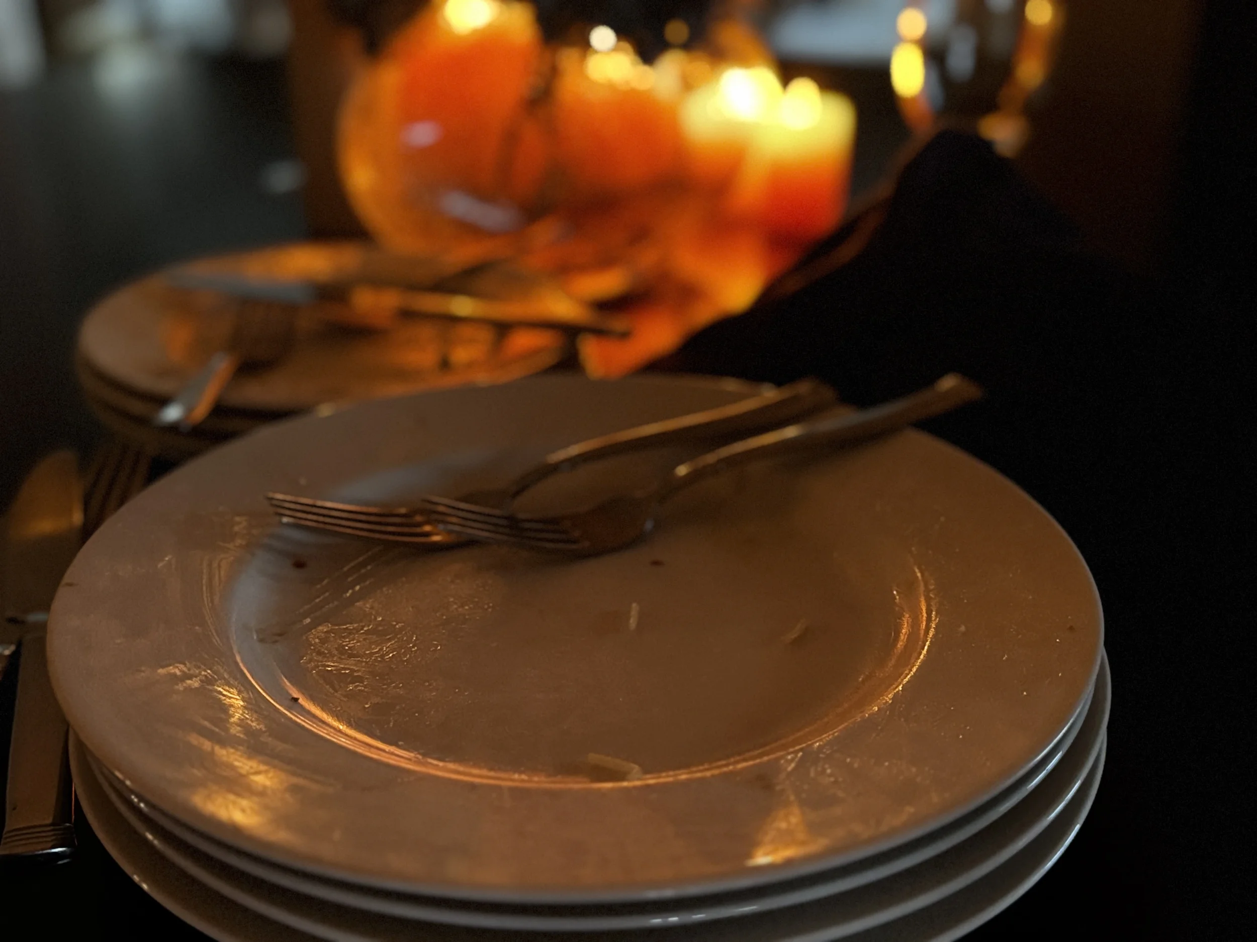 Empty dinner plates stacked after a shared meal, warm candlelight glowing in the background — the table after everyone has eaten, capturing the connection that builds in the space between the meal and goodbye.
