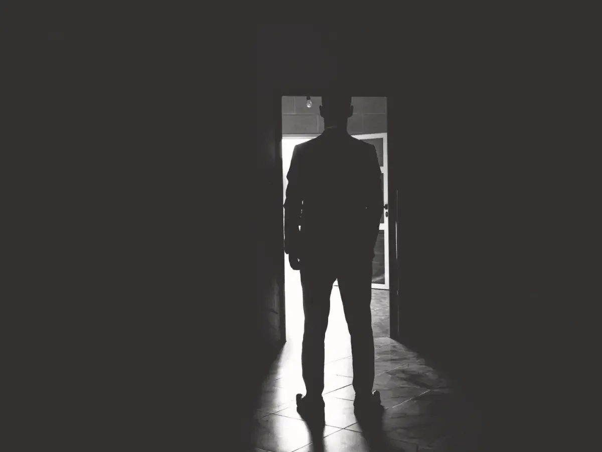 A silhouetted figure stands at a lit doorway in black and white, caught between darkness and light — opening and closing image for She The King's essay "When the Villain Feels Familiar," on the characters we're not supposed to relate to.