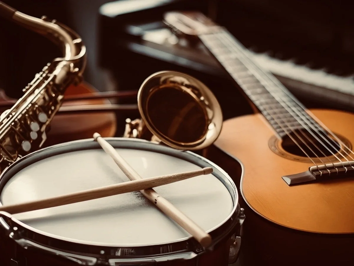 A collection of musical instruments including a saxophone, acoustic guitar, snare drum with crossed drumsticks, and a brass horn, arranged together in warm light.