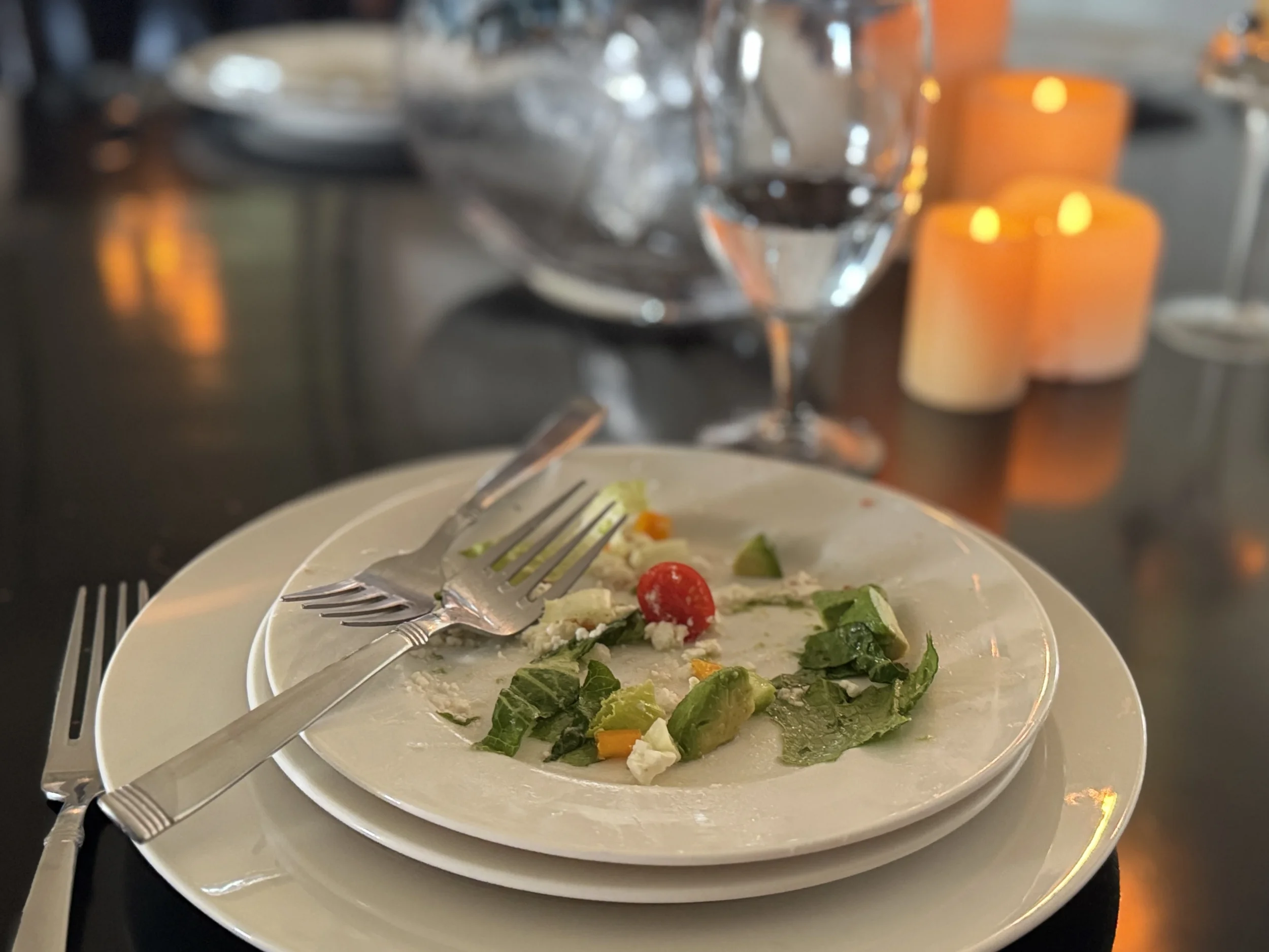 A nearly finished salad plate with a fork resting on it, wine glass and glowing candles blurred in the background — the feeling after a shared meal, illustrating the emotional connection built at the table.