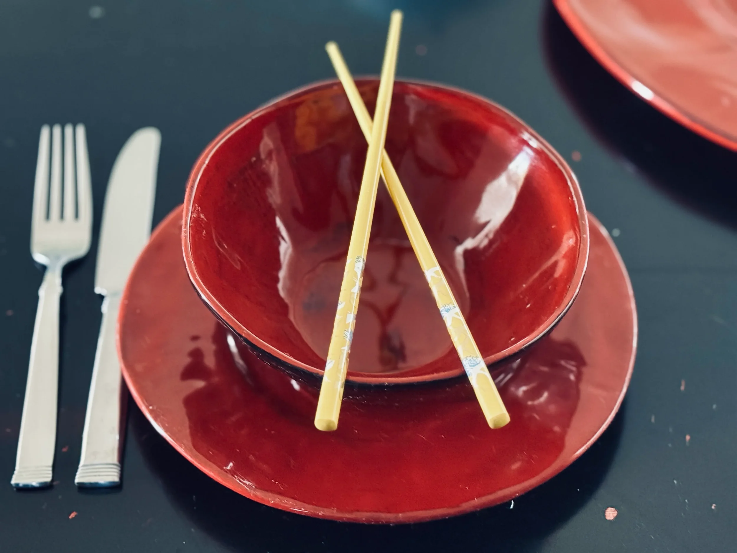 Empty red lacquer bowl with yellow chopsticks resting inside, placed alongside a Western fork and knife on a dark table, symbolising the Korean-English cultural in-between in Ada's story in Minbak