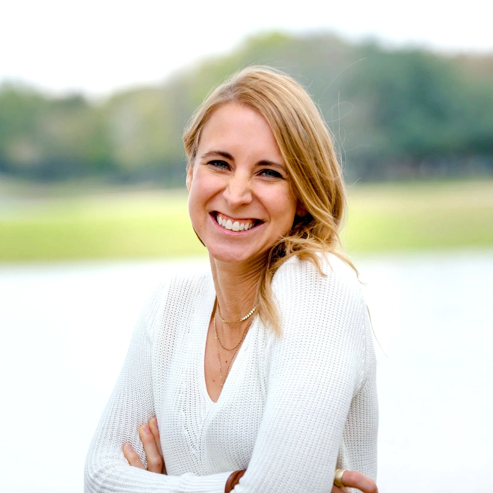 A woman with blonde hair wearing a white long-sleeve top standing outdoors near a body of water, smiling at the camera with trees in the background.