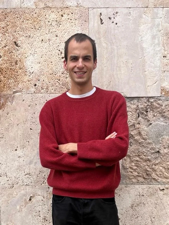 A guy standing against a wall, smiling, in a red sweater.