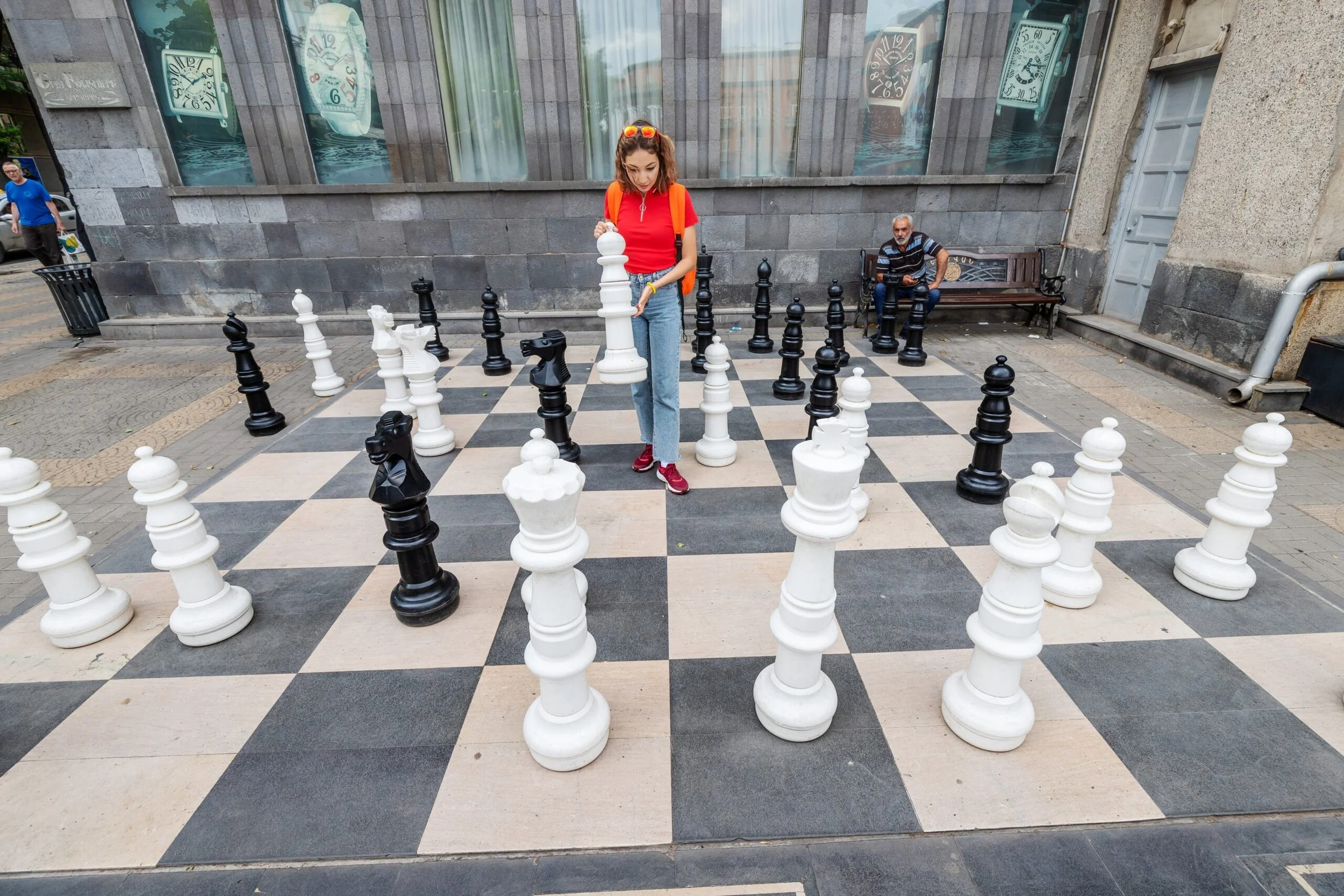 A woman playing chess on a large outdoor chessboard in a city street, with an older man sitting on a bench in the background.
