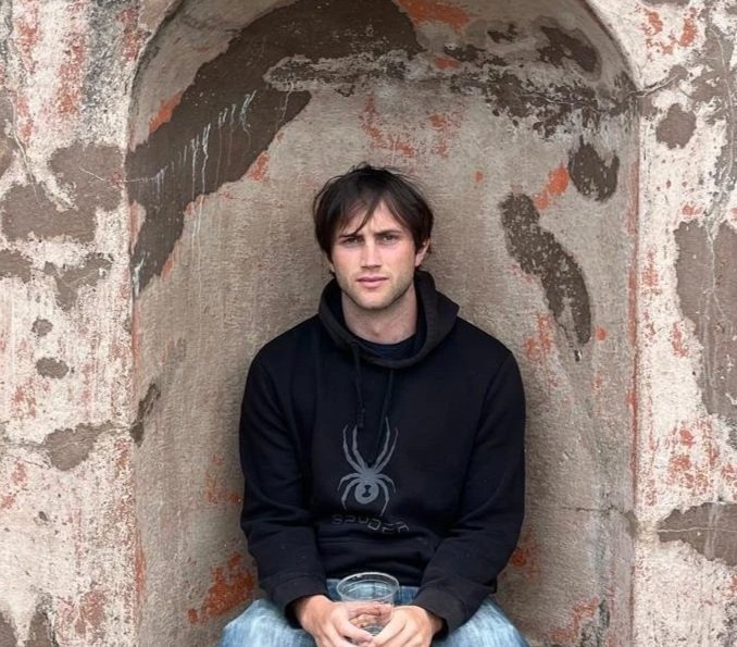 Young man with brown hair sitting in a brick nook, wearing a black hoodie and holding a glass of water.