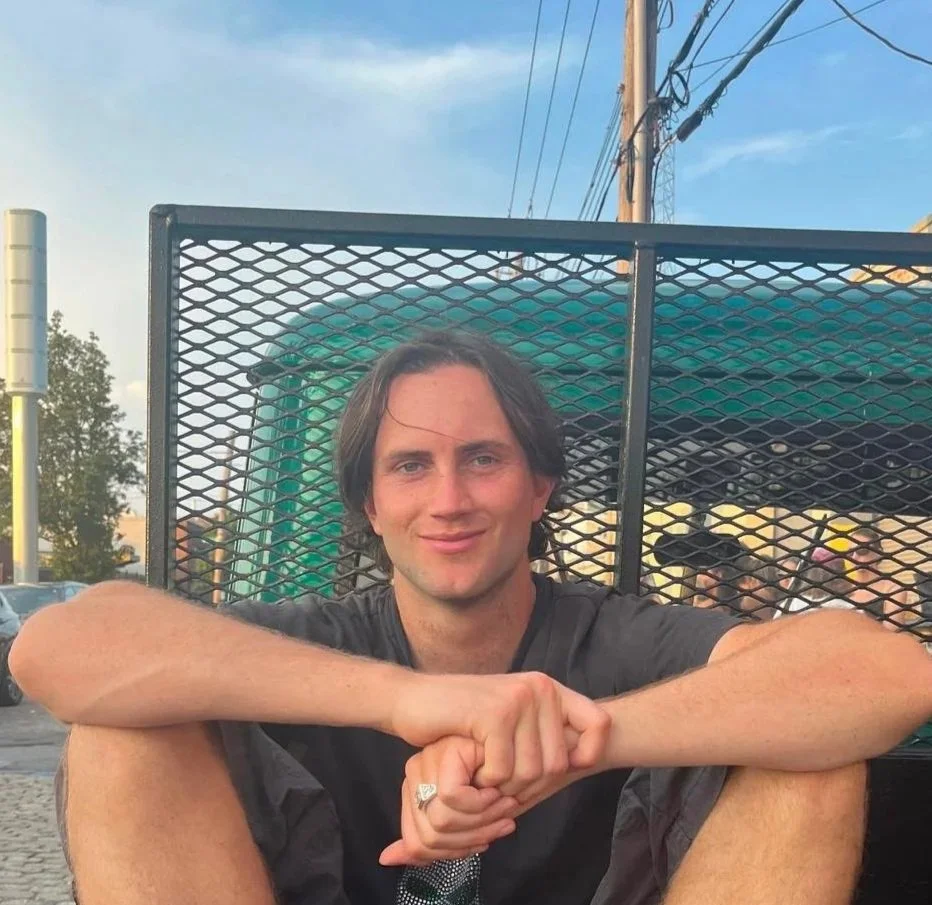 A young man with medium-length dark hair sitting outdoors, smiling and leaning on his knees with his arms crossed. He is wearing a dark T-shirt and is in front of a metal fence with a green structure behind it. The sky is partly cloudy with some power lines overhead.