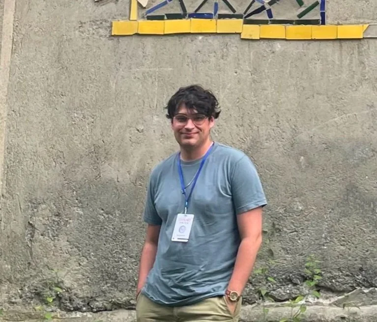 Young man with curly dark hair, wearing glasses, a light blue T-shirt, and a conference badge on blue lanyard, standing in front of a textured concrete wall with colorful decorative elements above.