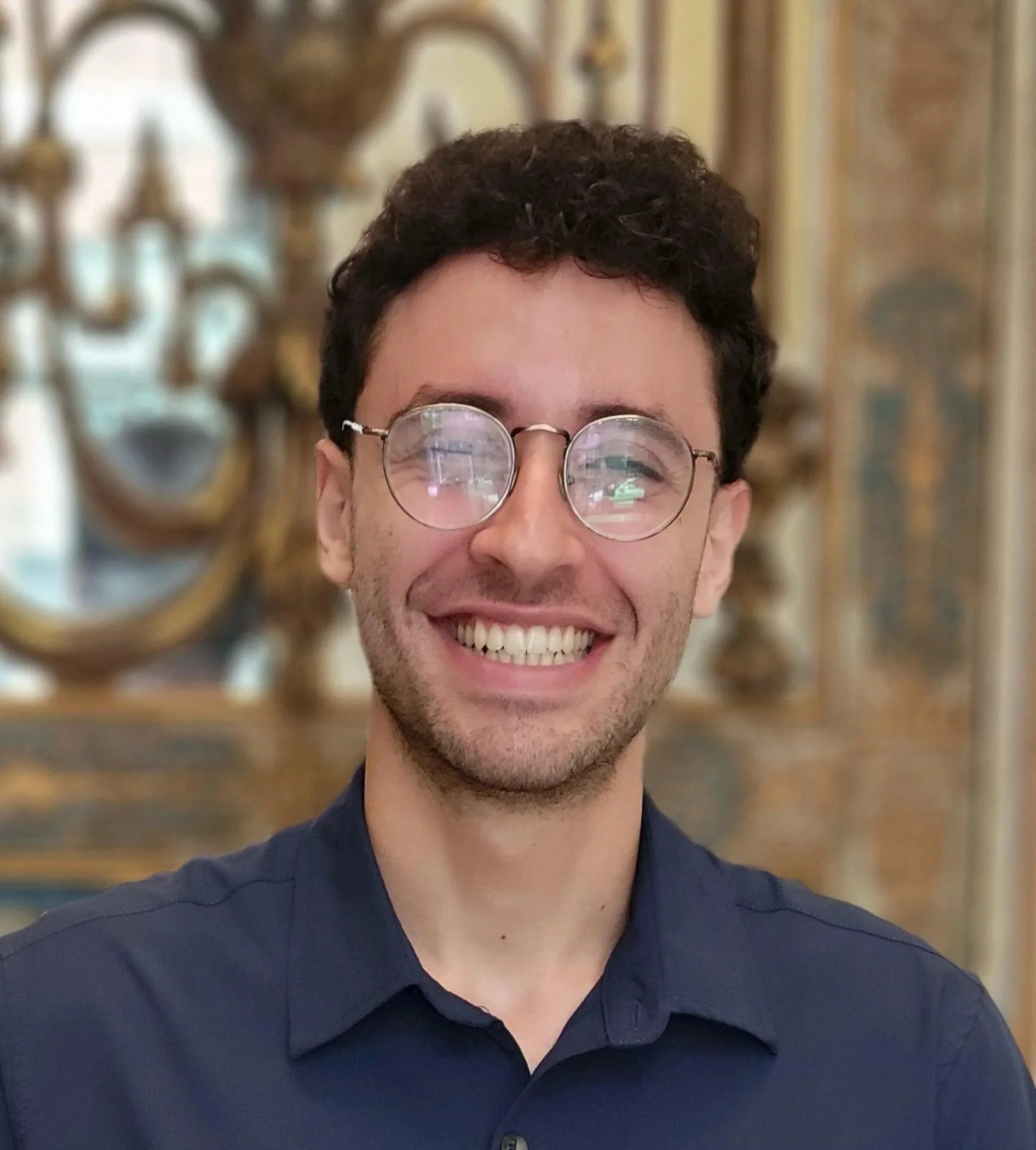 A young man with curly brown hair, wearing glasses and a dark blue shirt, smiling in front of an ornate gold and wood background.