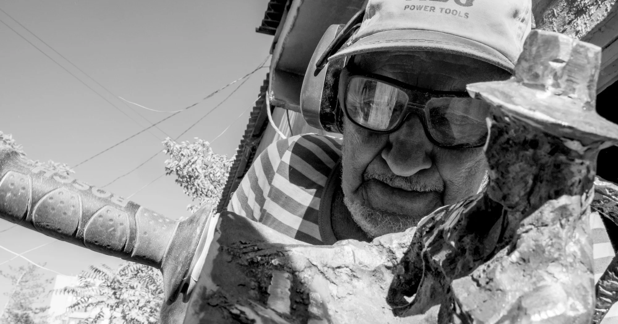 Black-and-white close-up of an older man in safety glasses and ear protection, working on a stone sculpture with a hammer or chisel.