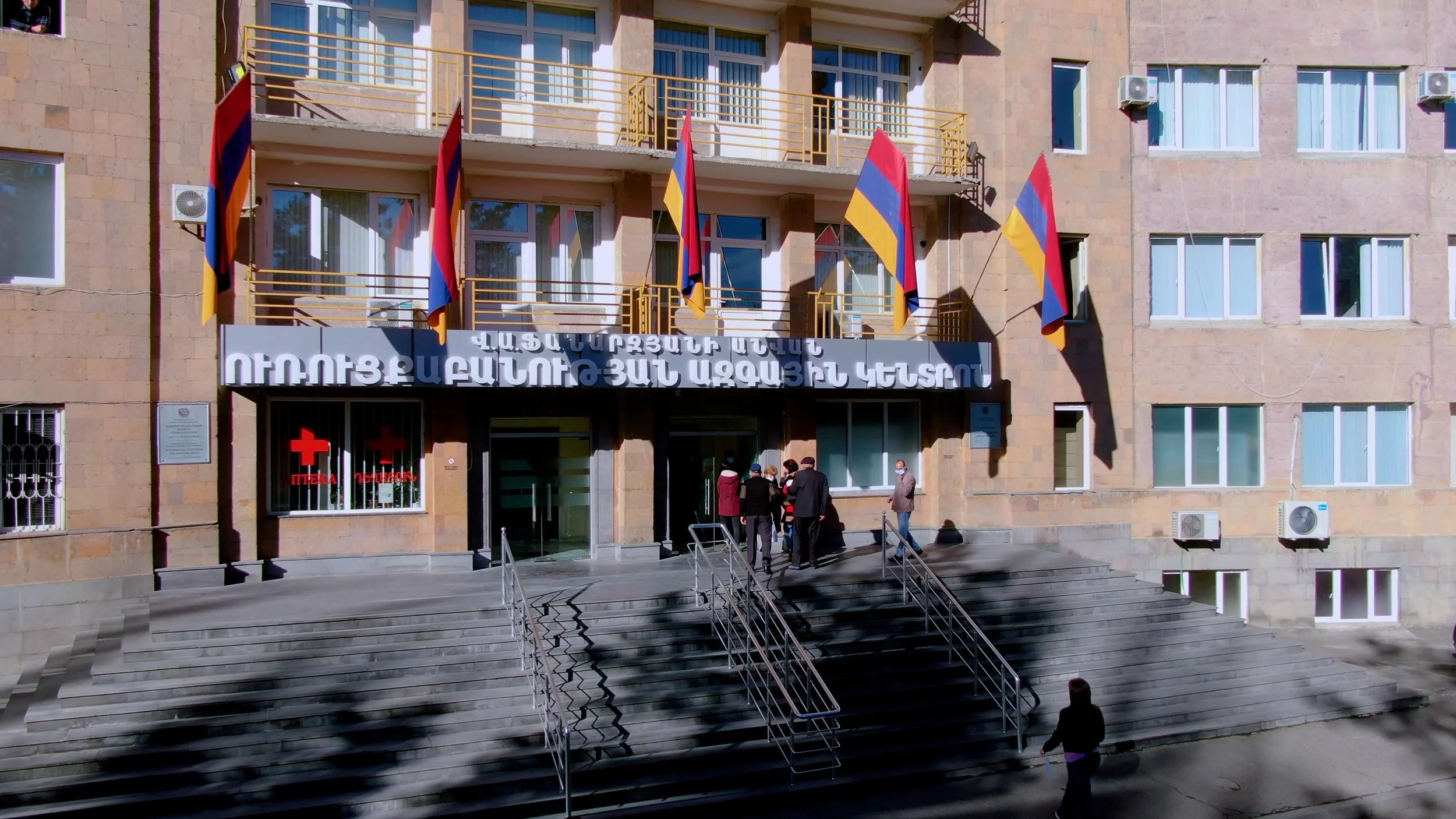 People gather at the entrance of the National Center of Oncology, decorated with Armenian flags.