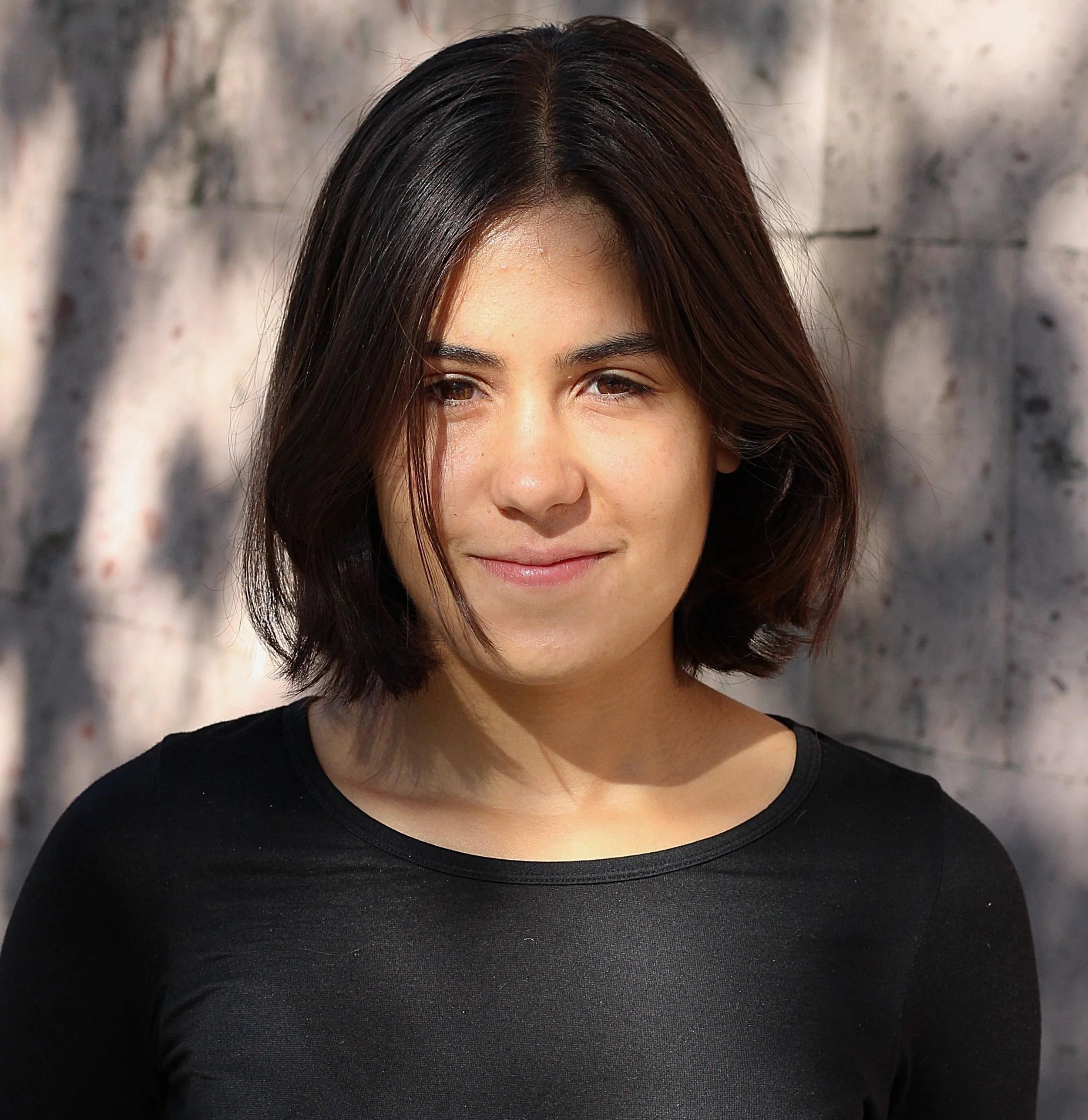 Close-up of a woman's face and hair against a gray stone background, wearing a black top.