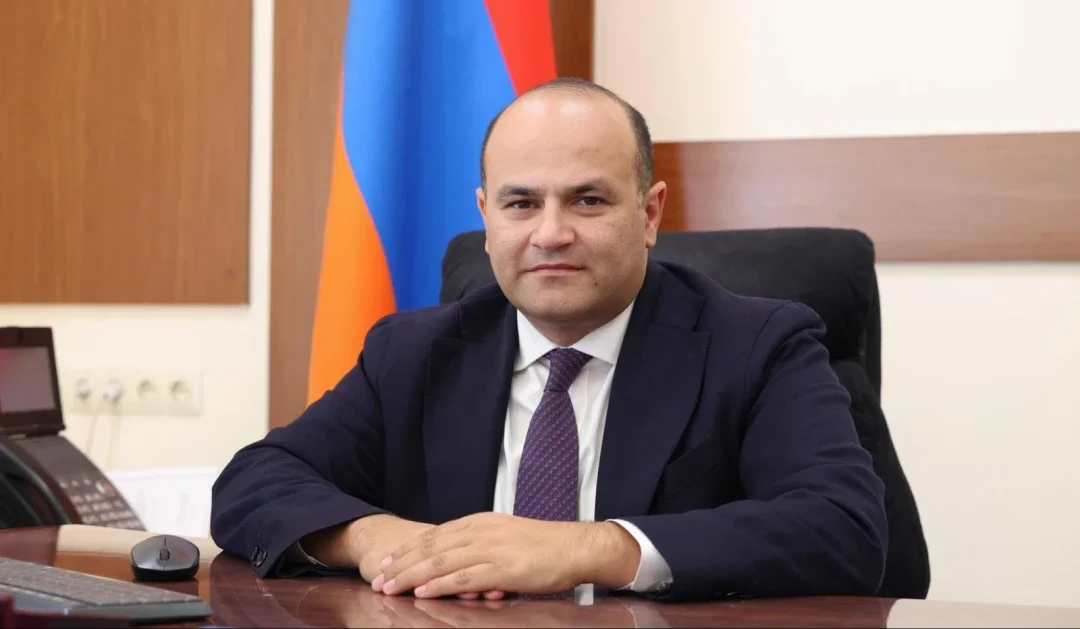 Armenia's Ambassador to the U.S. Narek Mkrtchyan sits at his desk in front of the Armenian flag, hands folded, wearing a dark suit and patterned tie.