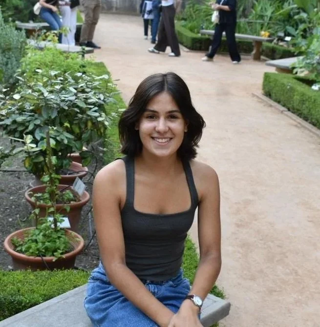 A young woman with shoulder-length dark hair, wearing a black tank top and blue jeans, sitting on a bench in a botanical garden or park with potted plants nearby.