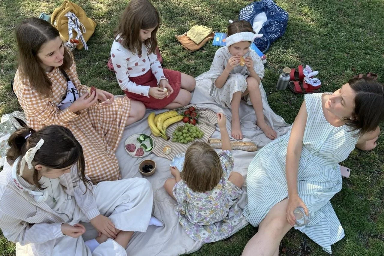 A group of children and an adult having a picnic on a blanket in a park. They are surrounded by food such as bananas, grapes, strawberries, and snacks. The children are eating and sitting on the blanket, some with headbands and casual clothes. The adult is sitting on the edge of the blanket with a jar in hand.