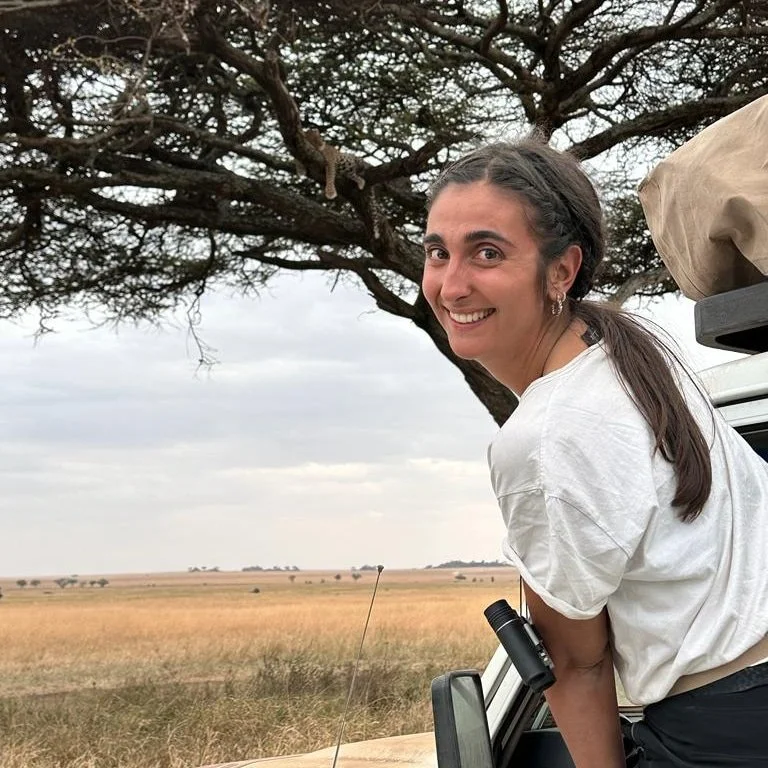 Young woman leaning out of a safari vehicle with binoculars, smiling, over grassland with scattered trees under a cloudy sky.