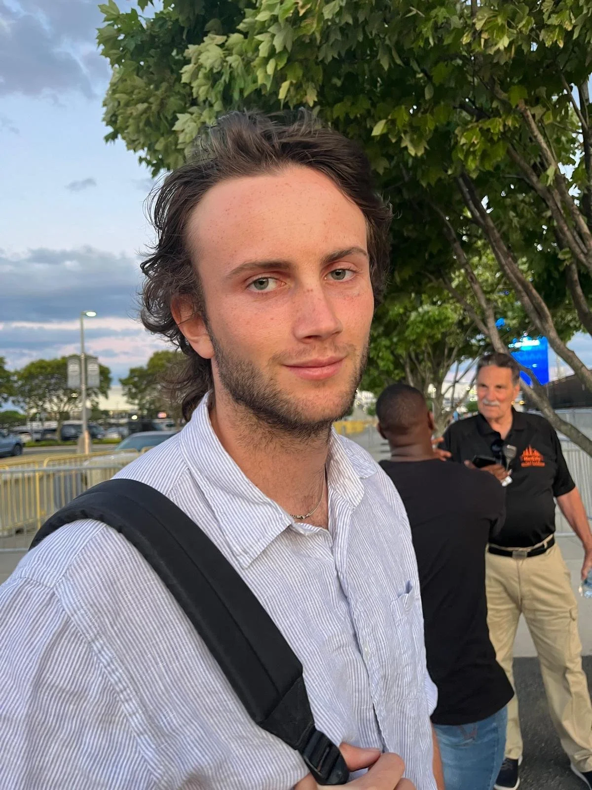 A young man with light skin and brown hair, wearing a light-colored striped shirt and a backpack, stands outdoors near green trees with a cloudy sky in the background. Two men are visible in the background, one wearing a black shirt and the other in a black polo with orange text.