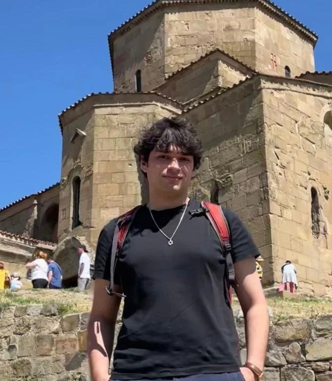 Young man with curly dark hair, wearing a black t-shirt and red backpack, standing in front of an old stone church with rounded towers and small windows, under a clear blue sky.