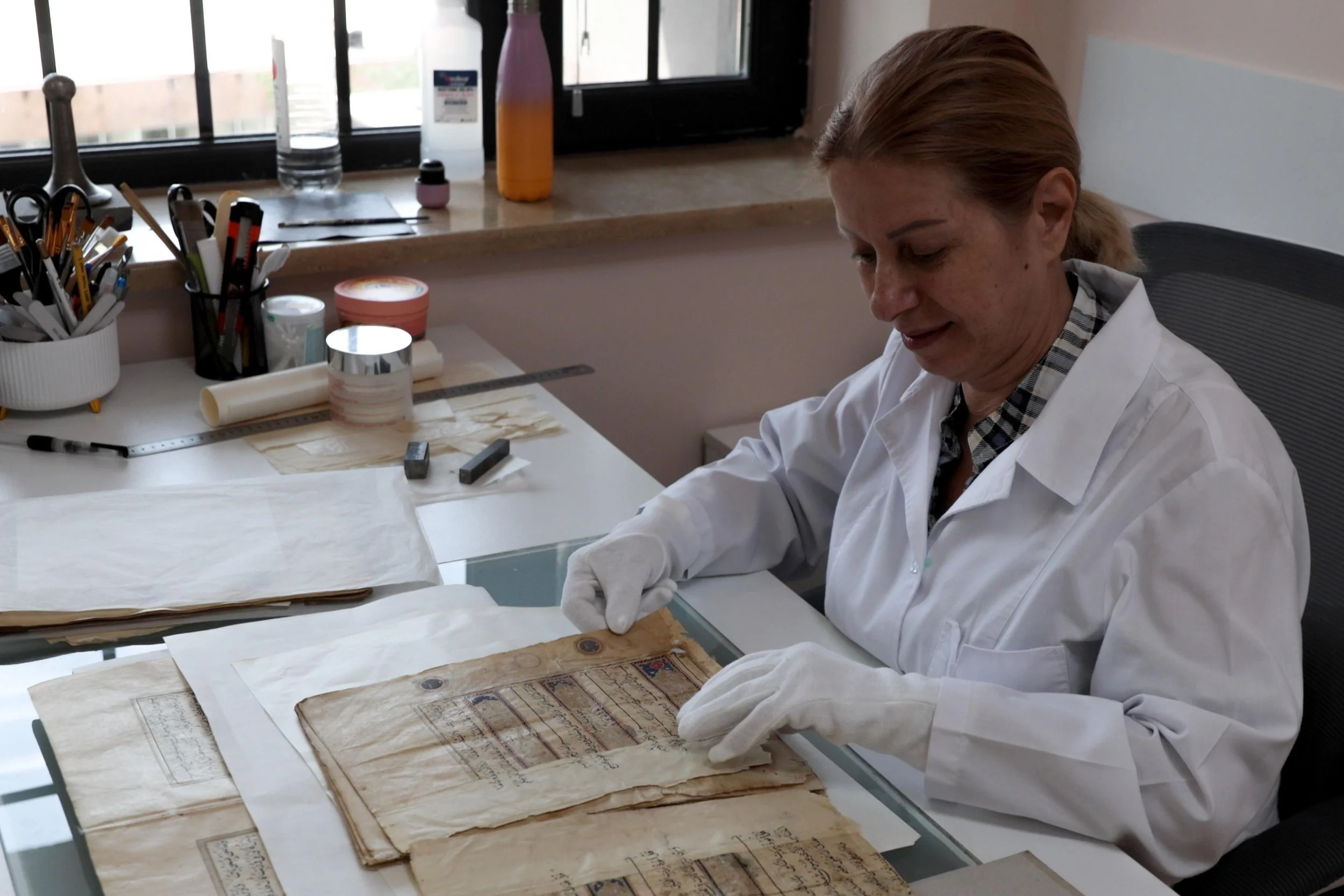 A gloved conservator examines an old illuminated manuscript page on a worktable surrounded by restoration tools.