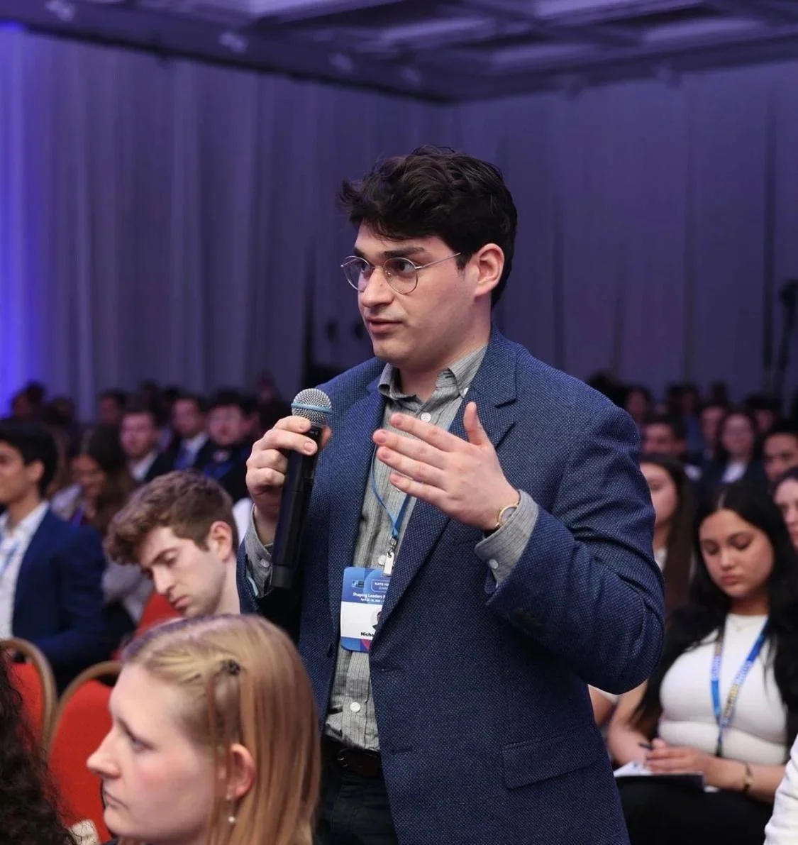 A young man with dark curly hair and glasses holding a microphone and speaking during a conference or seminar, with a seated audience in the background.