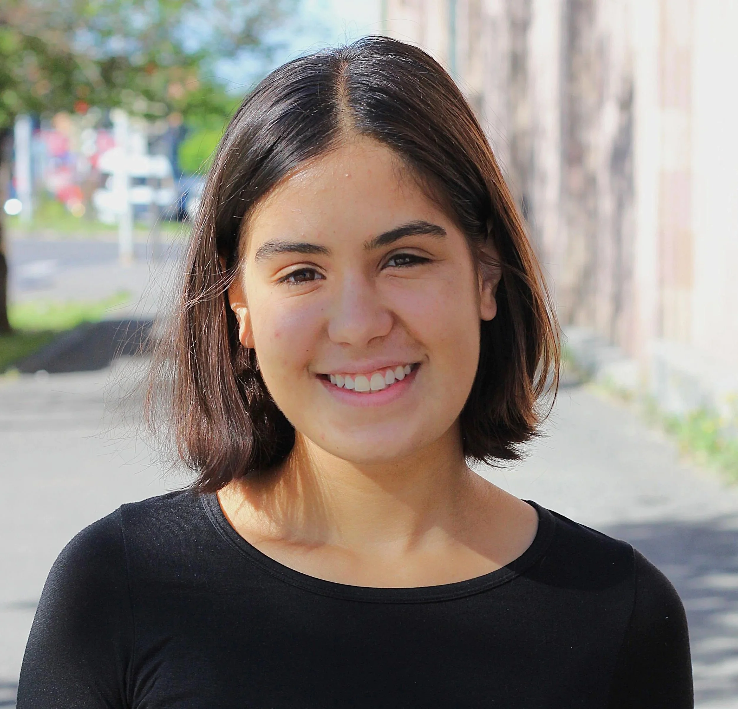 Close-up of a woman's face smiling, with shoulder-length brown hair, wearing a black top, outdoors with blurred trees and street in the background.