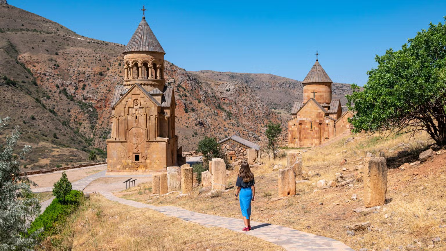 A woman walking on a paved path towards two historic stone churches with conical roofs, set in a dry, mountainous landscape under a clear blue sky.