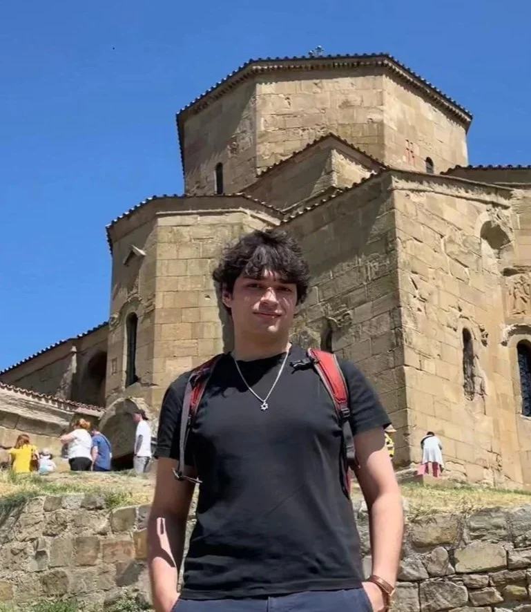 A young man wearing a black t-shirt and backpack standing in front of an historic stone church on a sunny day.
