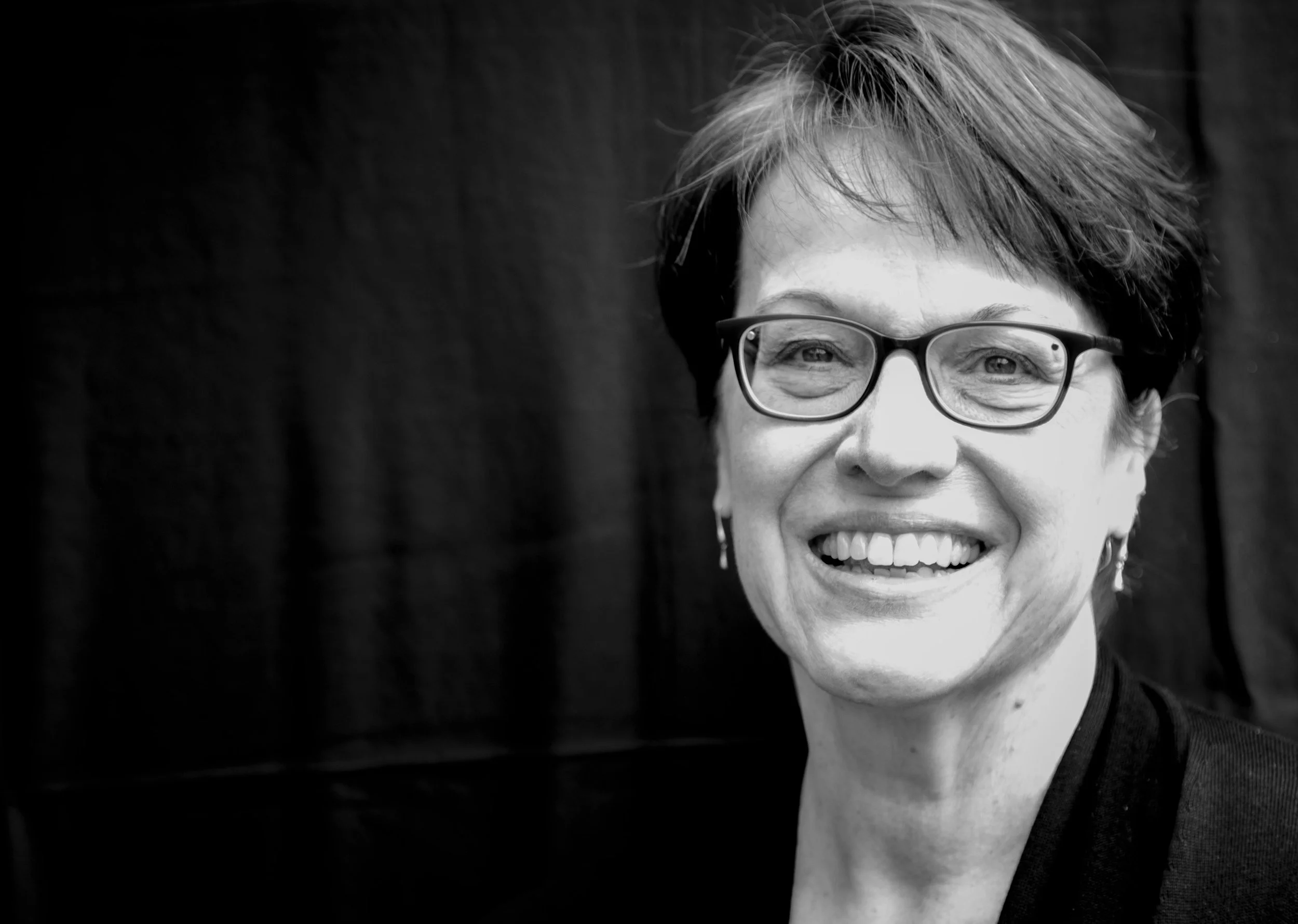 Patty Brandmaier, a white woman with short hair and glasses smiling in front of a dark backdrop