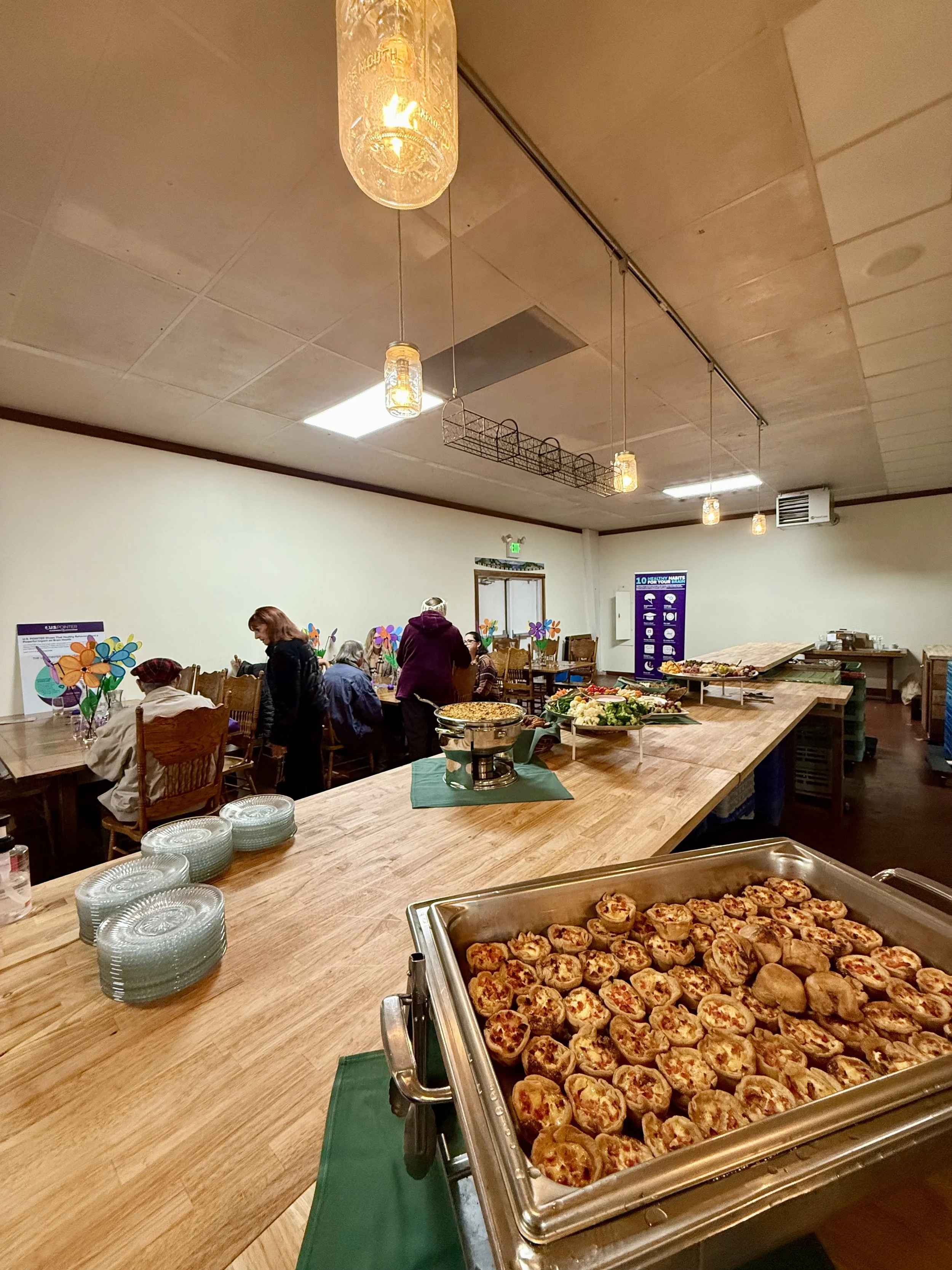 Buffet setup with pizza rolls in a chafing dish, plates stacked on the counter, and a group of people dining in the background in a room decorated with colorful paper flowers.