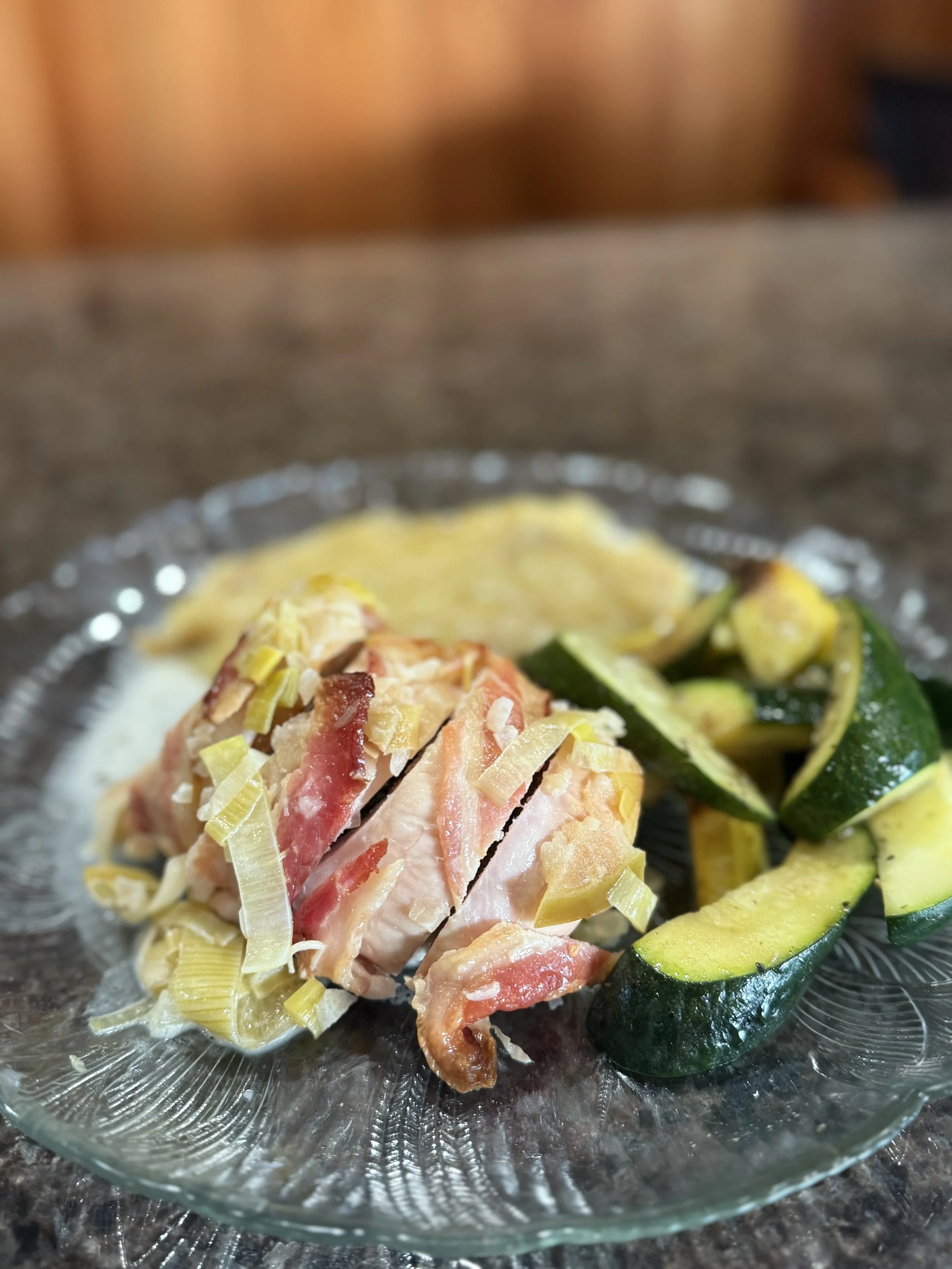 A glass plate with cooked chicken topped with sliced leeks, served with cooked zucchini on a dark countertop.