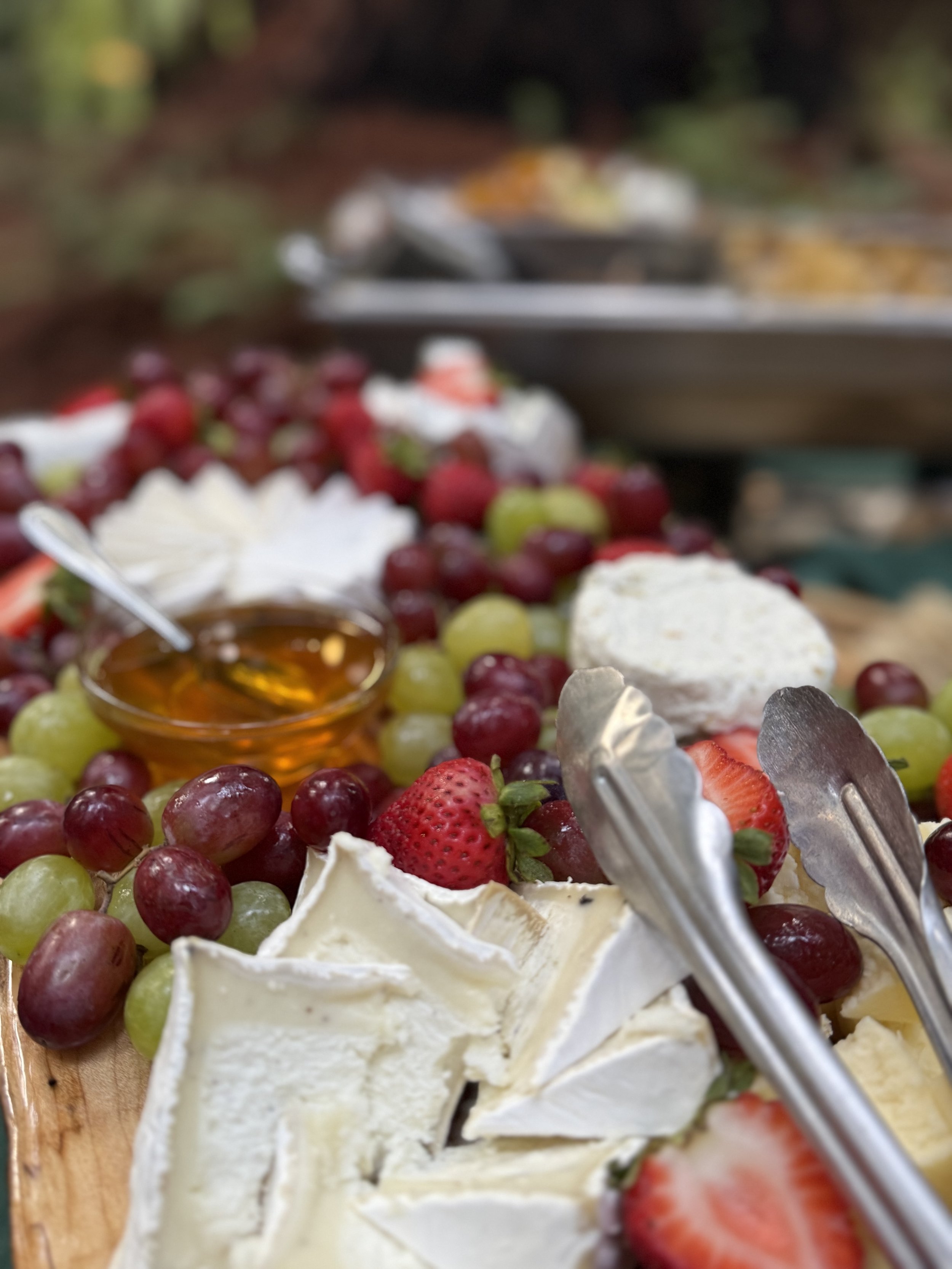 Close-up of a cheese and fruit platter with strawberries, grapes, and honey, with serving tongs in focus, at an outdoor setting.