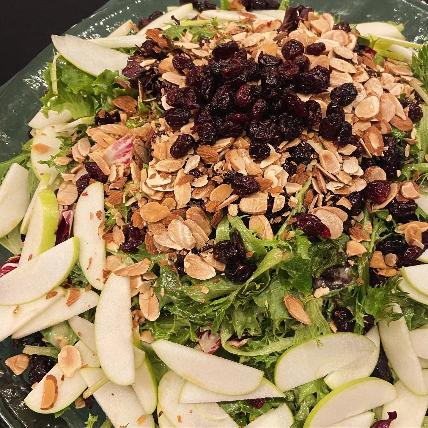 A fresh salad with sliced apples, greens, dried cranberries, and chopped nuts, arranged on a glass plate.