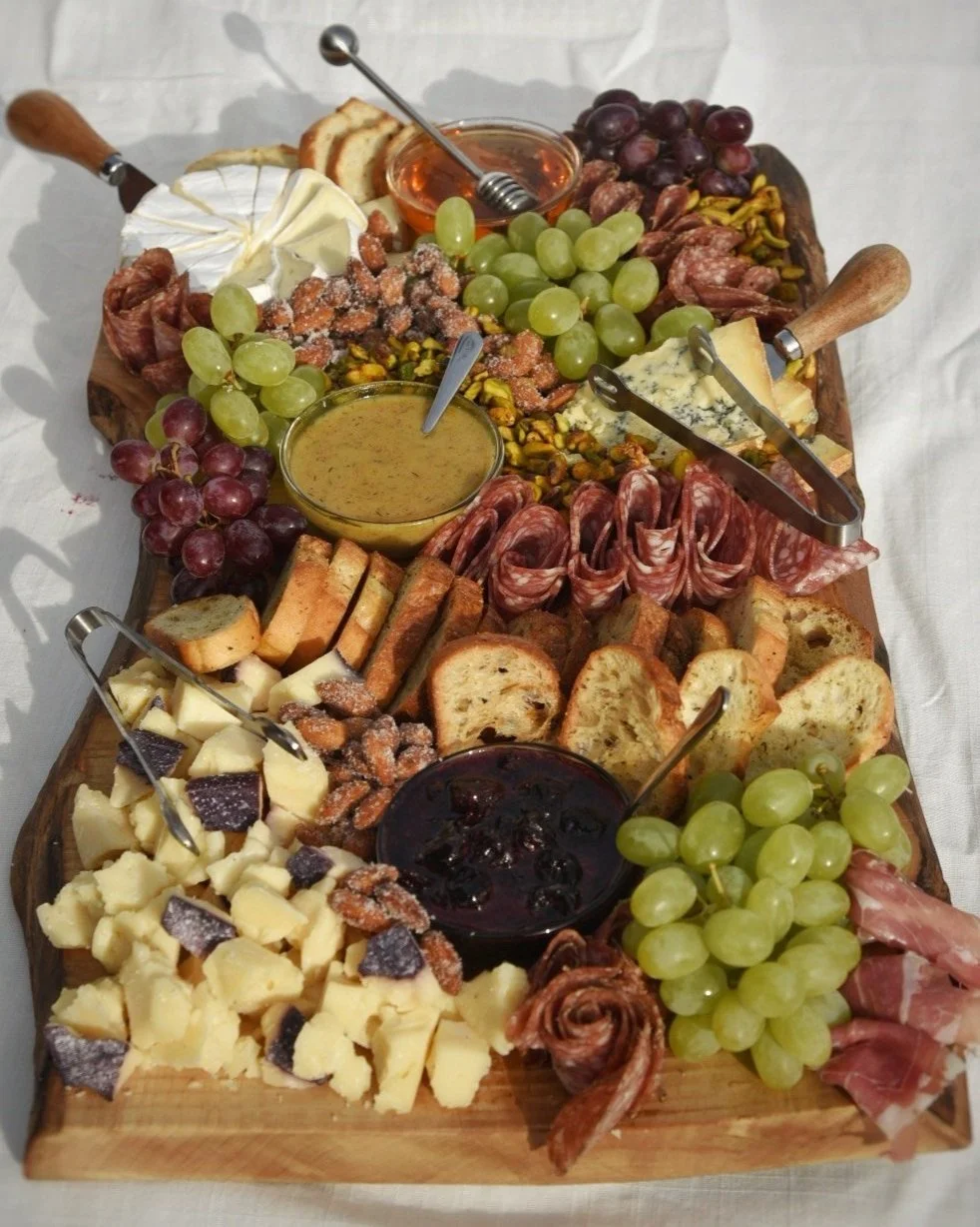 A wooden cheese and charcuterie board with various cheeses, grapes, crackers, nuts, salami, and jams.