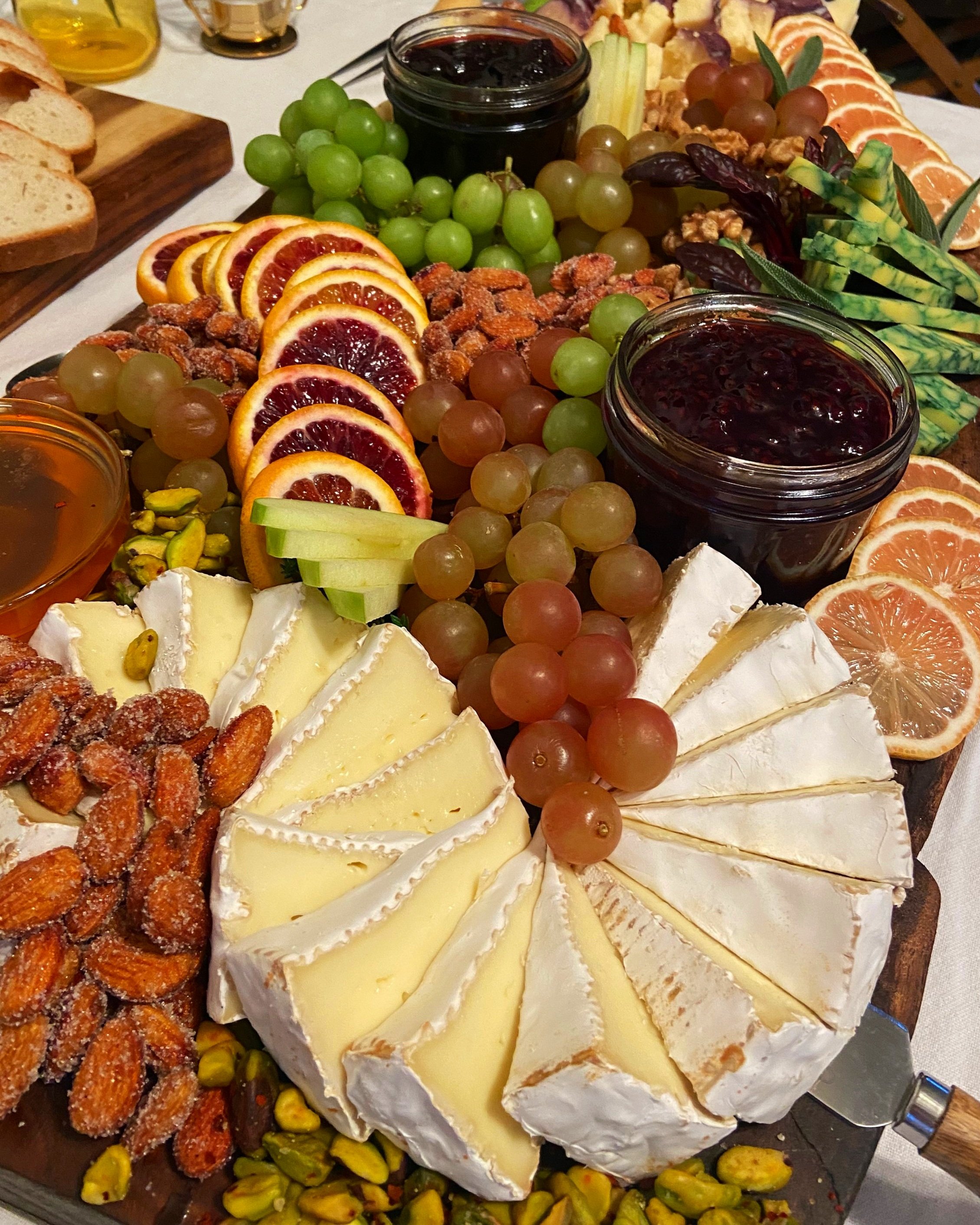 A cheese platter with brie, grapes, blood orange slices, and assorted crackers, with clusters of green and purple grapes, drizzled honey, and jars of jam.