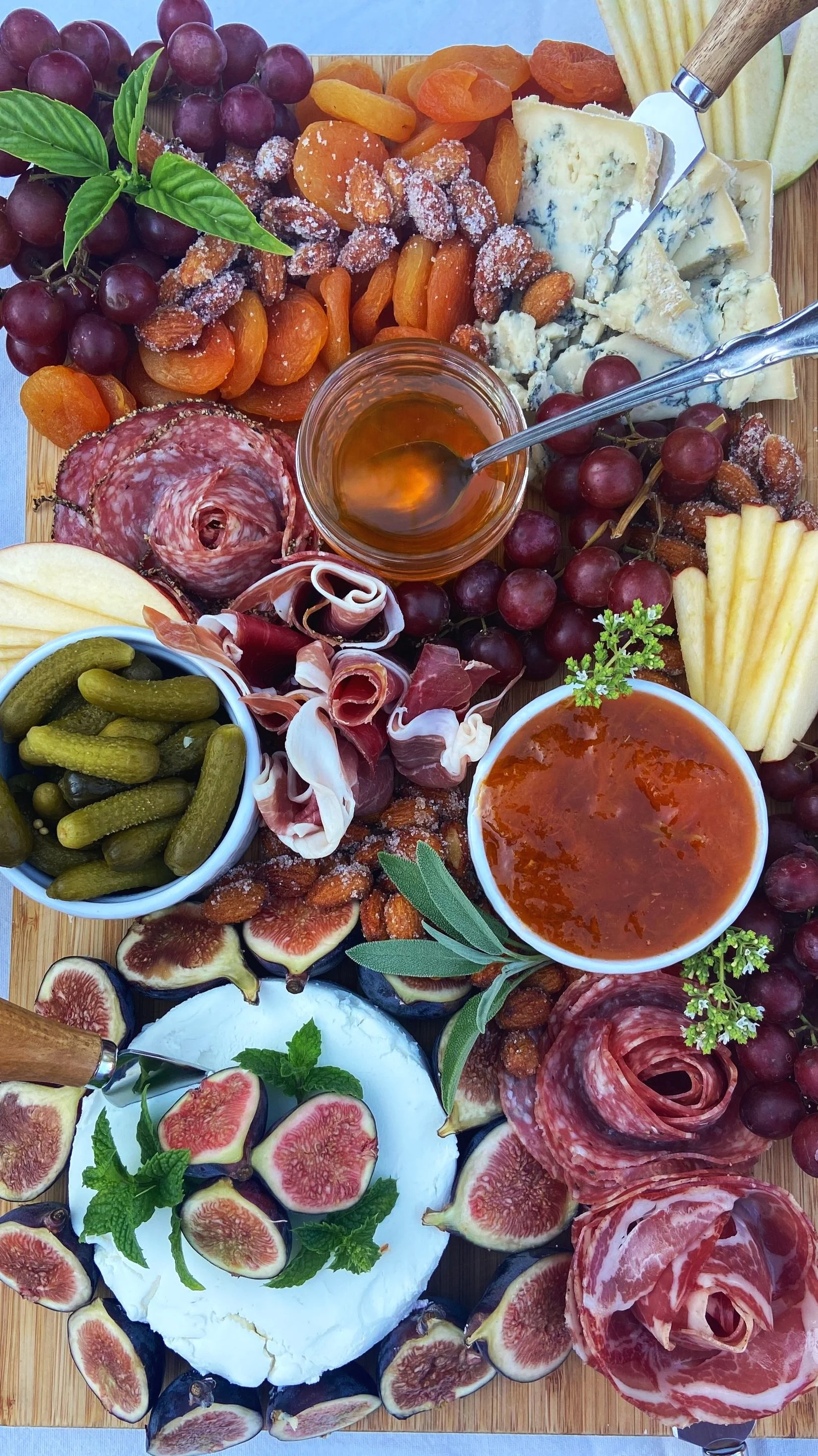 A cheese and charcuterie platter with grapes, dried apricots, almonds, figs, honey, dips, crackers, and fresh herbs.
