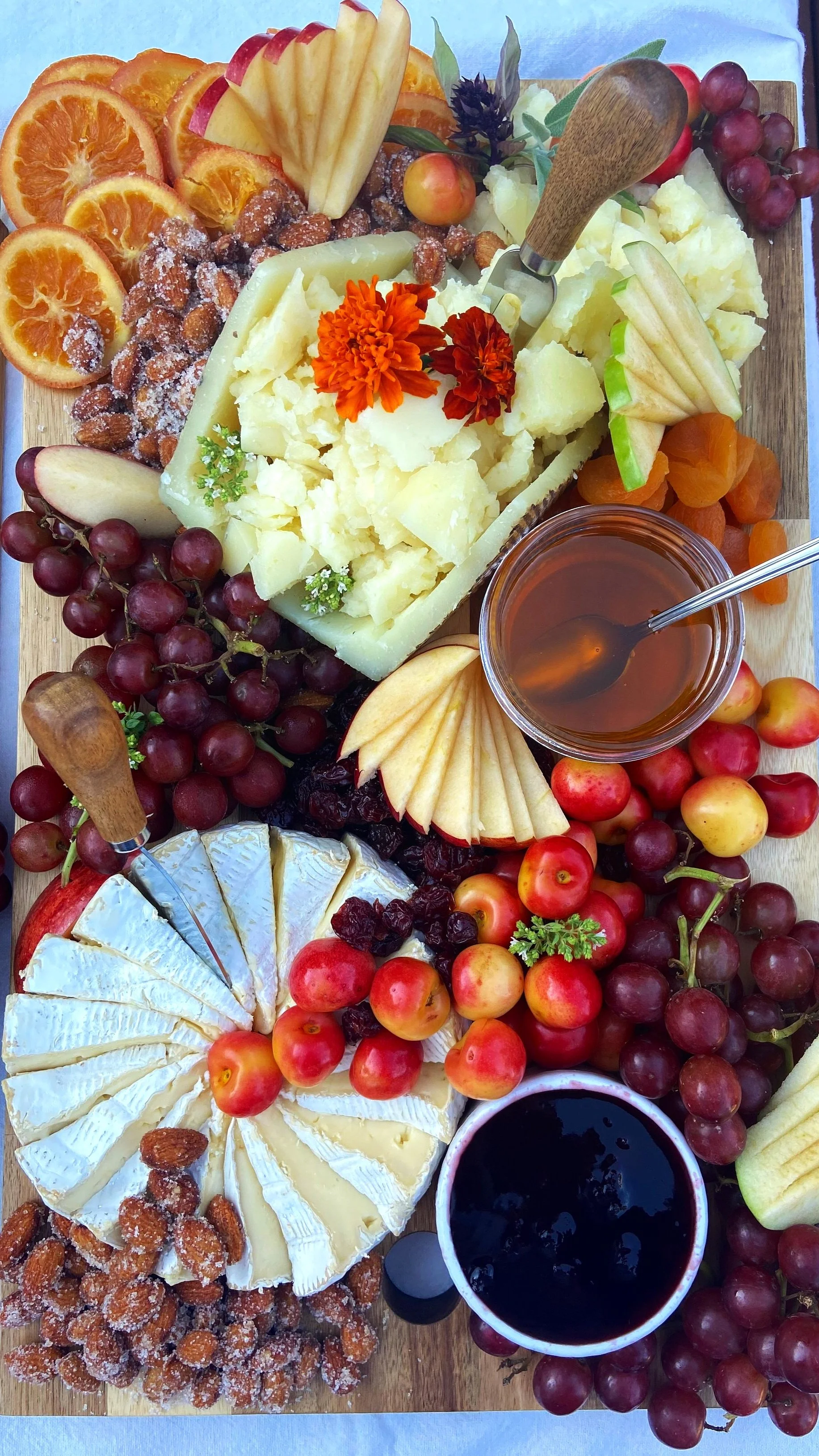 A cheese board with assorted cheeses, fresh fruit including grapes, apples, and dried fruit, honey, and flowers.