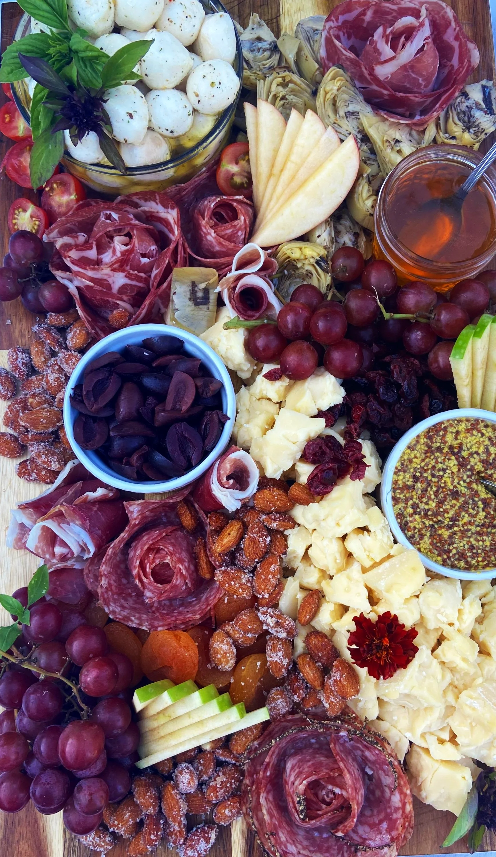 Charcuterie board with cheese, grapes, cured meats, nuts, fruit, honey, and garnishes.
