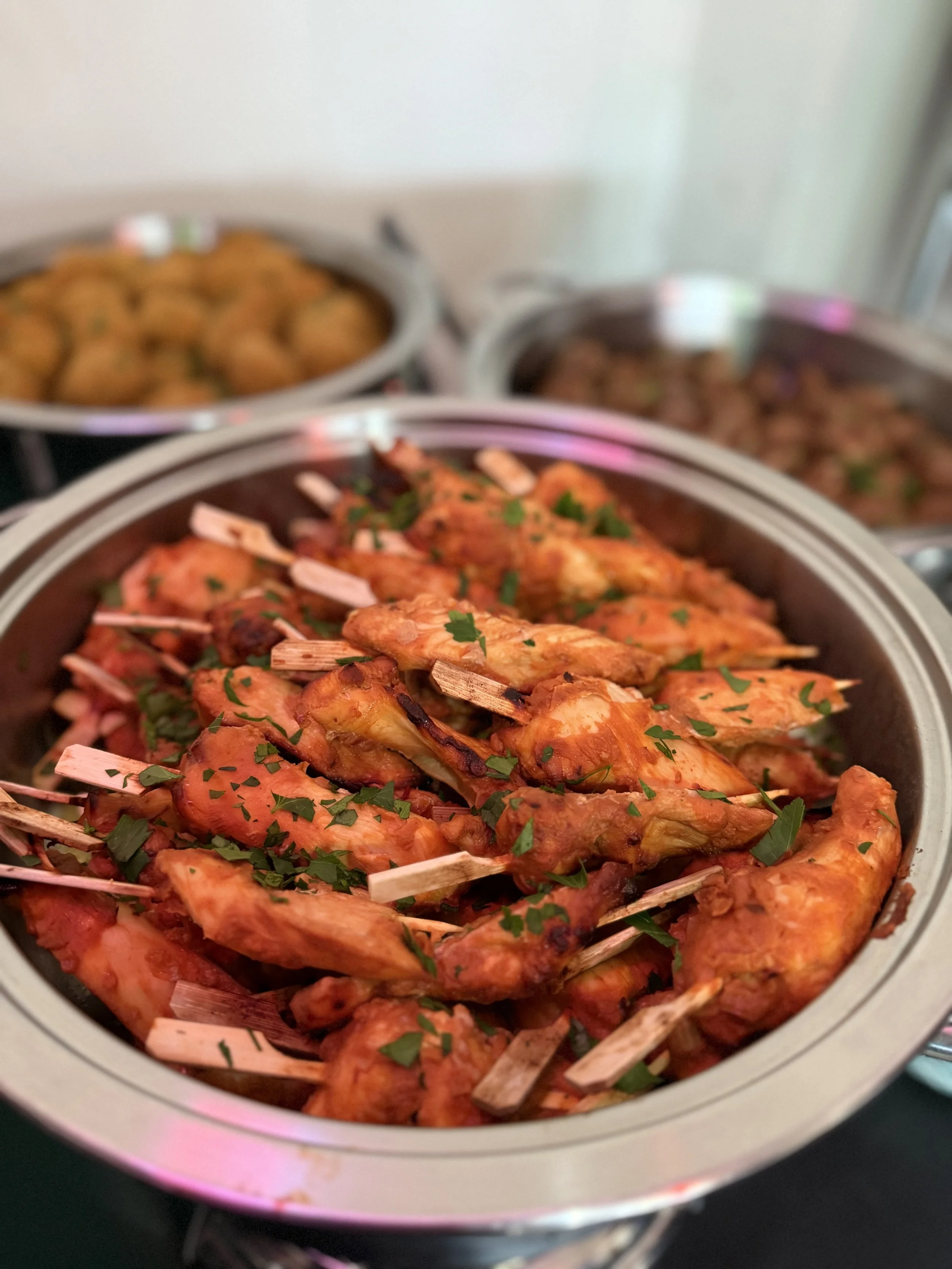 Chafing dish filled with grilled chicken skewers garnished with chopped herbs, with more dishes in the background.