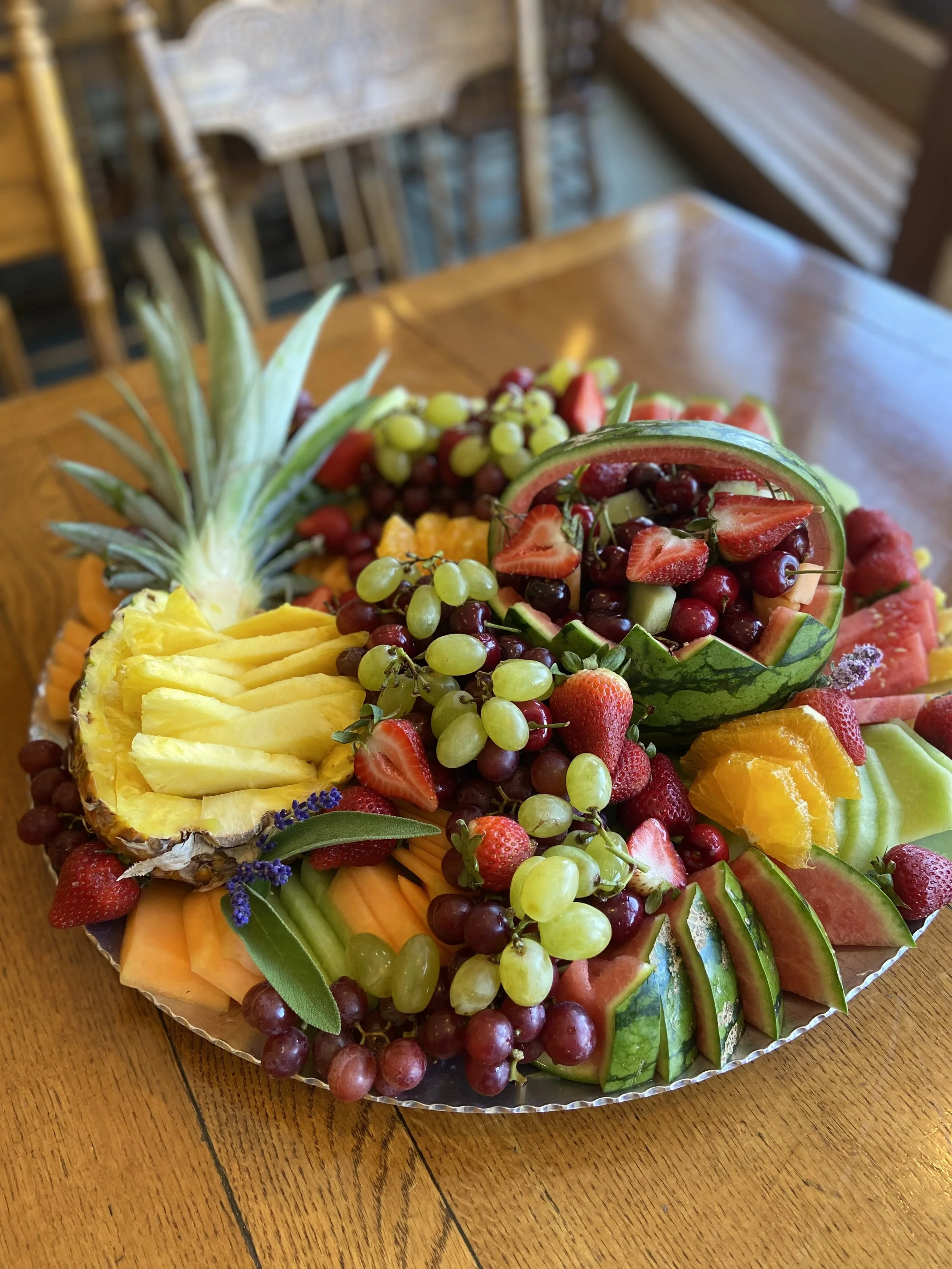 Plate of assorted cut and whole fruits including pineapple, watermelon, strawberries, grapes, cantaloupe, honeydew, oranges, and a pineapple top, on a wooden table.