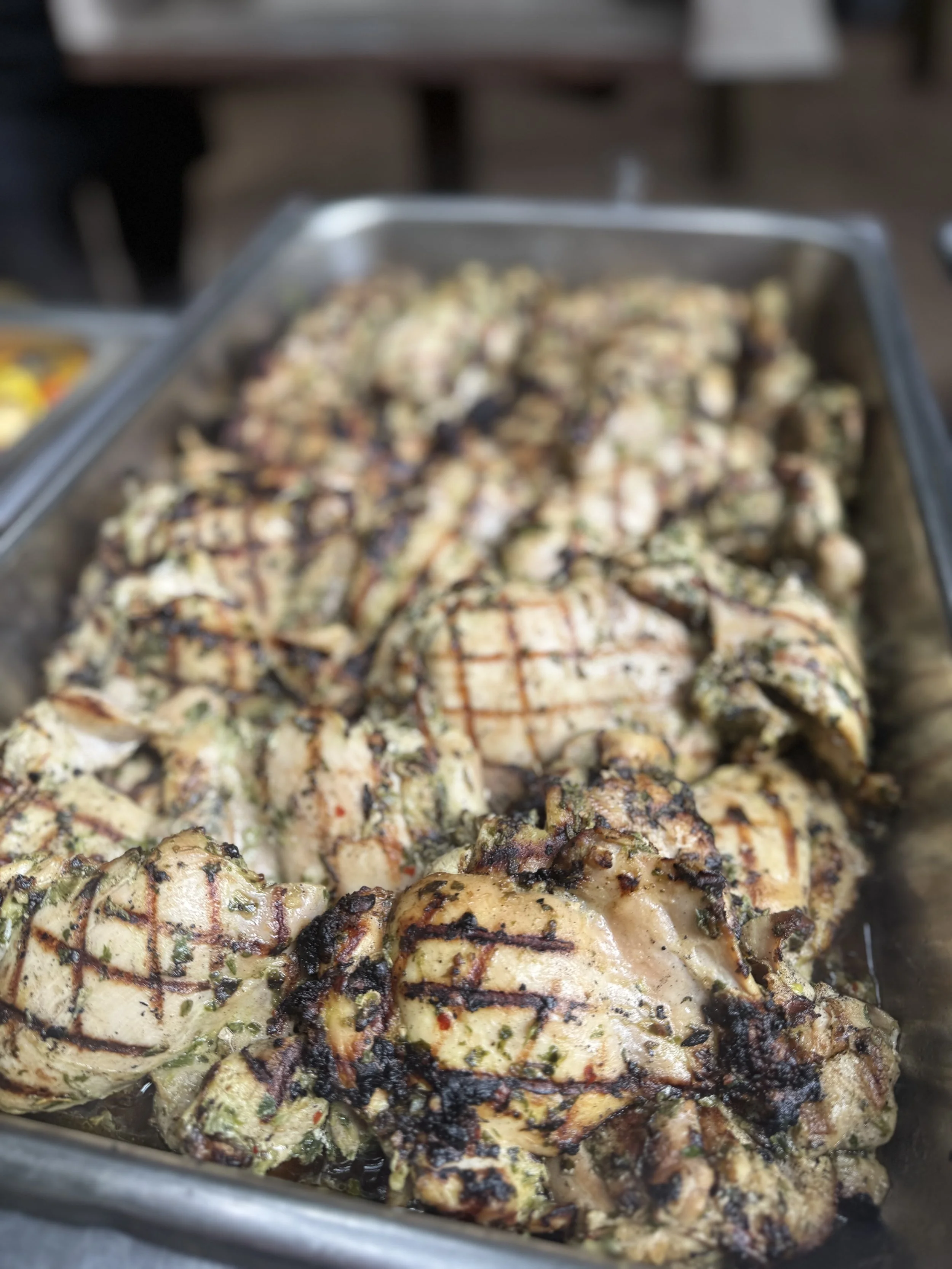 Grilled chicken pieces in a stainless steel tray with grill marks and herbs.