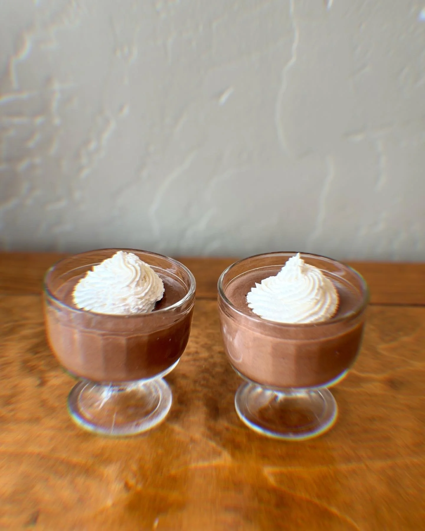 Two glasses of chocolate mousse with whipped cream topping on a wooden surface.