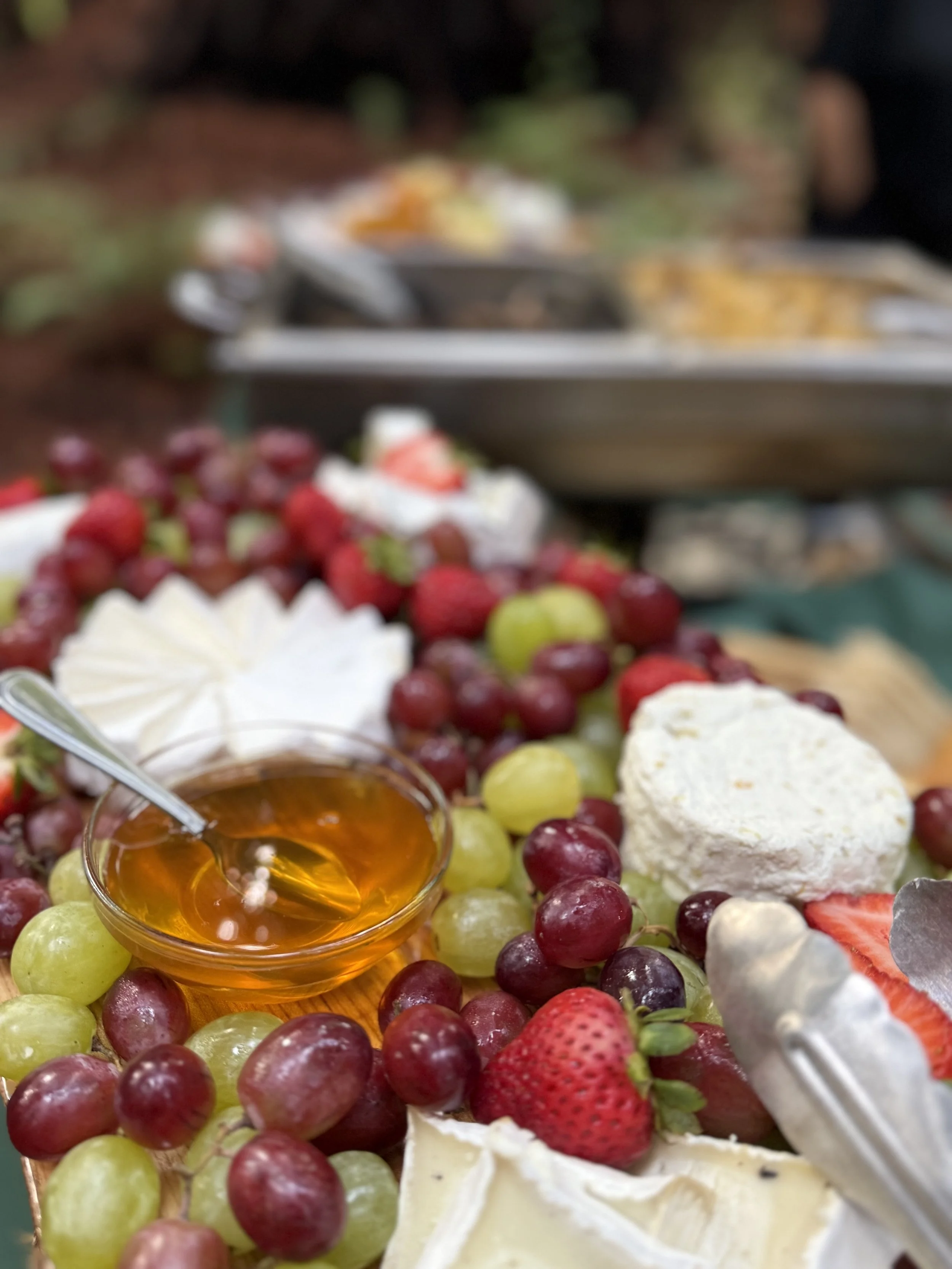 A platter of fresh grapes, strawberries, a wheel of cheese, and honey, with additional food items blurred in the background.