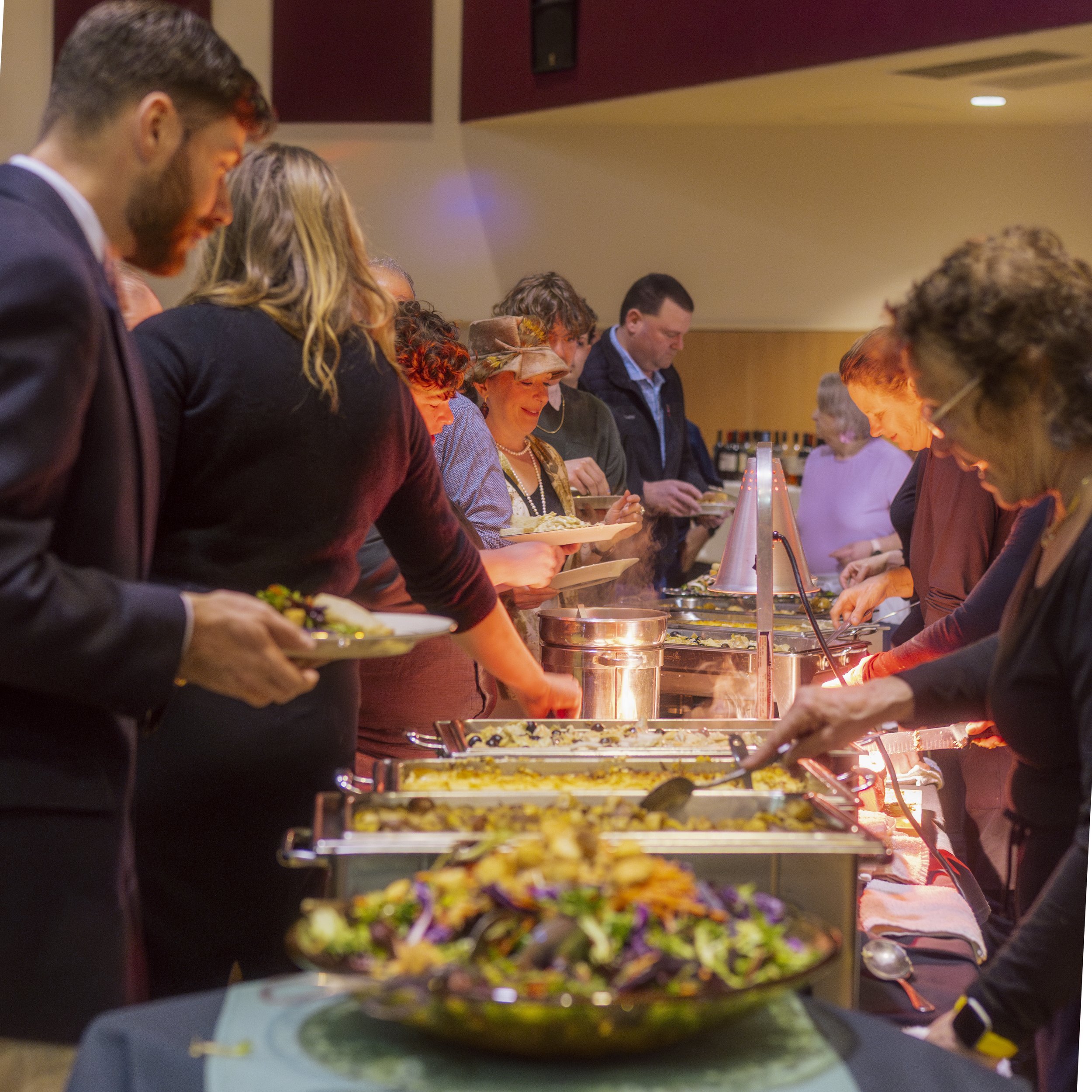 People serving themselves food at a buffet table in an indoor setting with warm lighting.